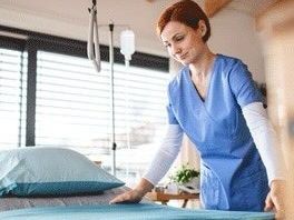 Nurse in blue scrubs making a bed in a hospital room.