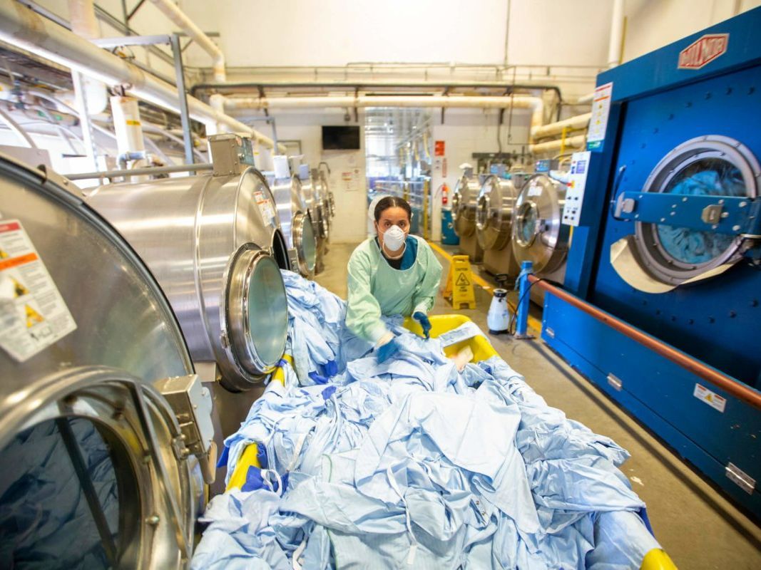 Laundry worker in PPE pushing a cart of linens in a commercial laundry facility with large washers.