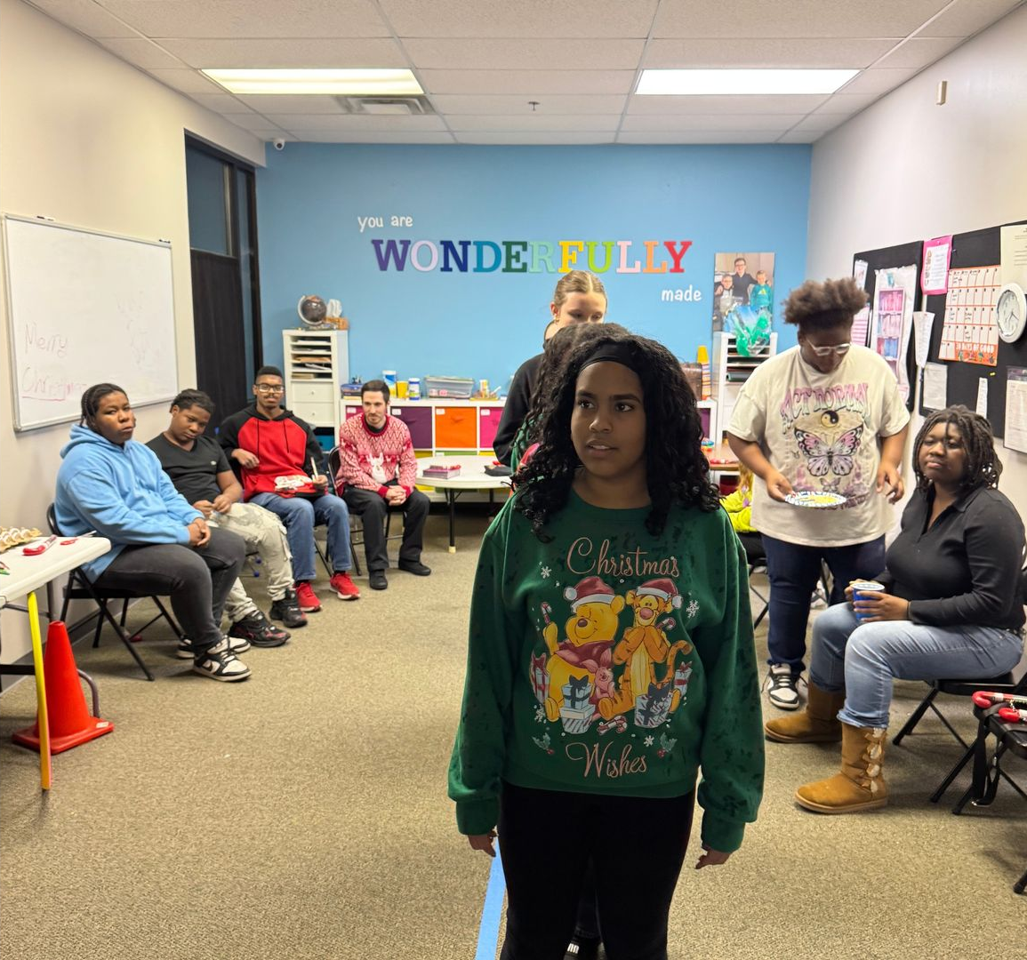 Child standing in a classroom, with adults seated around the room and a “Wonderfully” mural on the back wall
