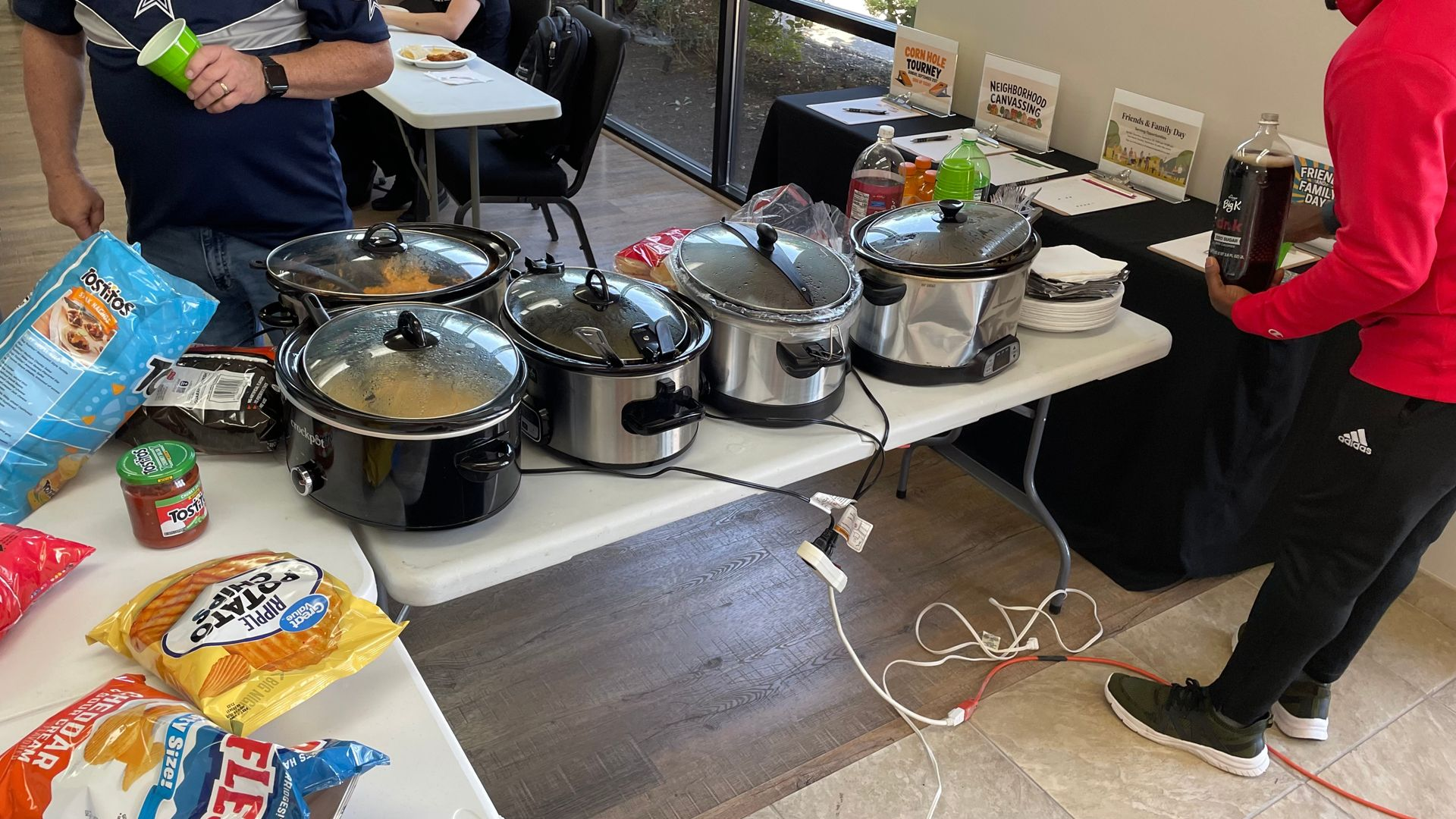 Catered buffet table with several slow cookers, snacks, and two people serving food indoors