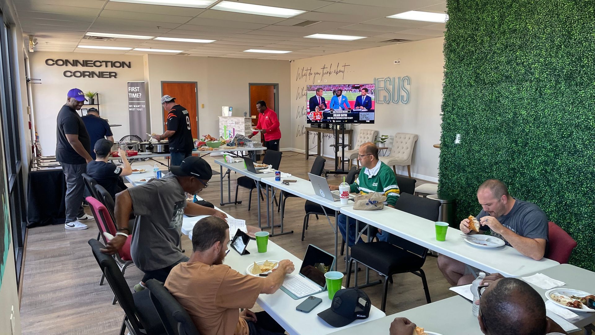 People seated at tables in a bright meeting room, watching a TV screen during a group event.