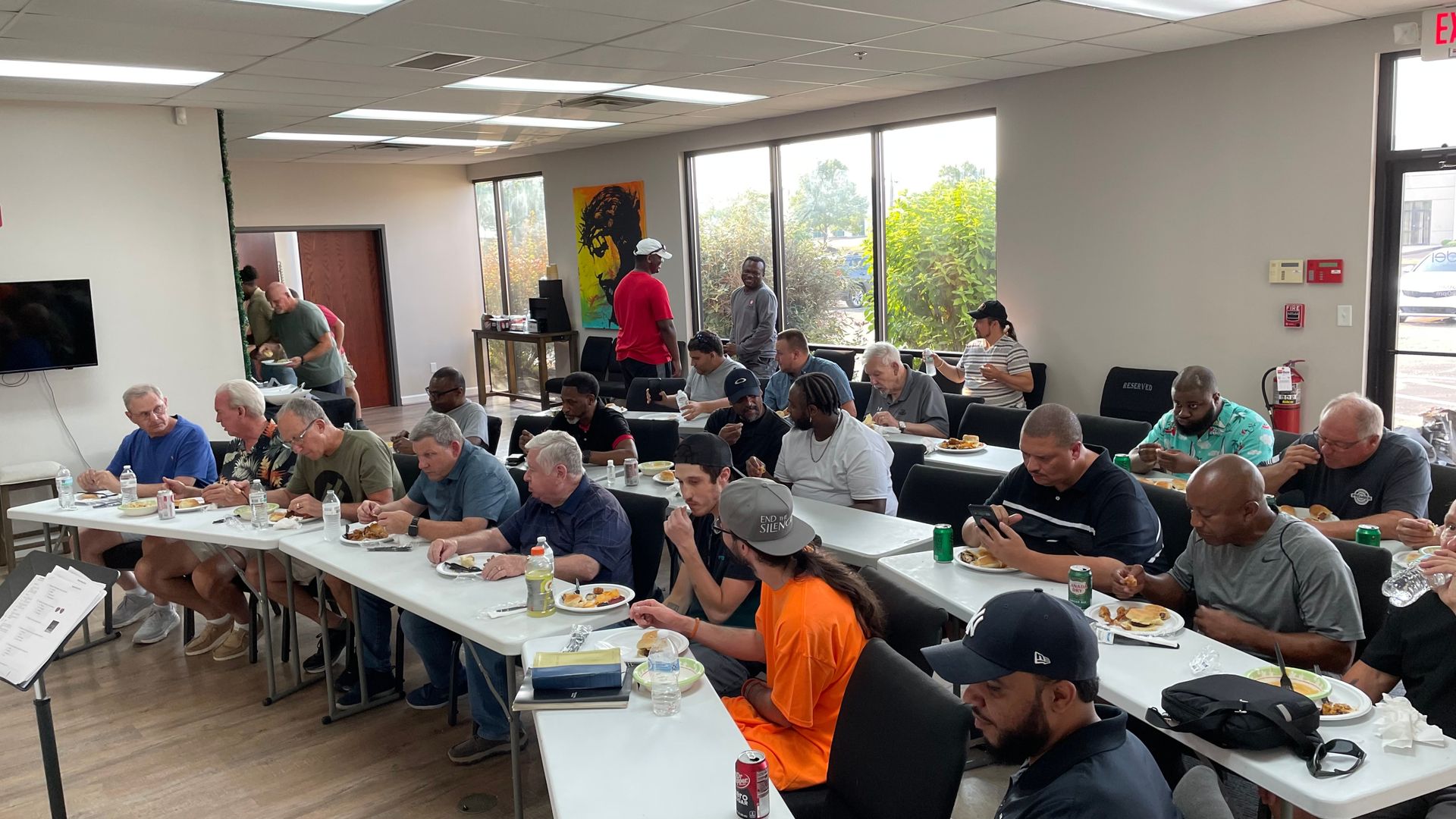 Group of people seated at long tables in a bright room, eating and talking during a community meal