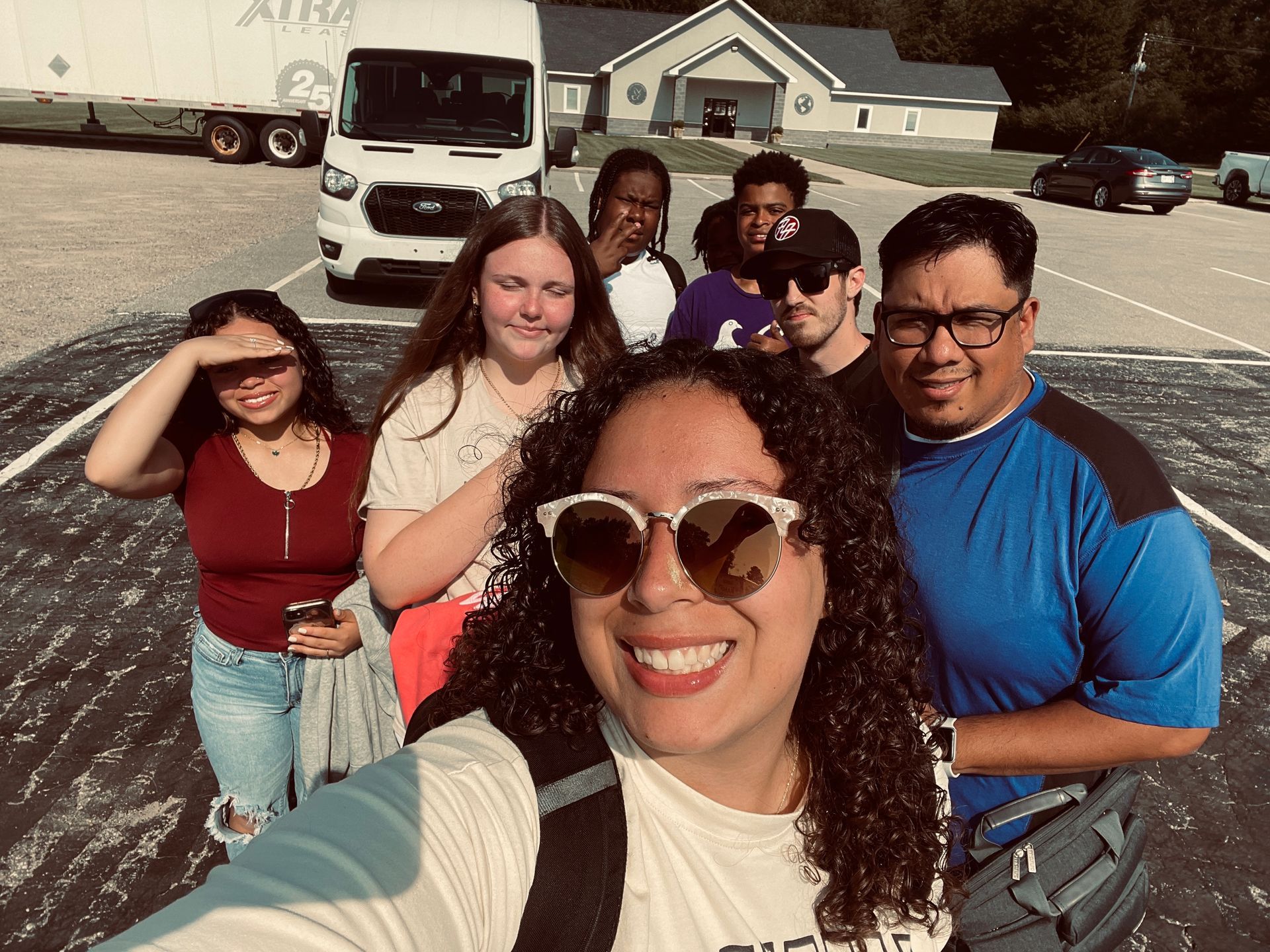 Group selfie of six people in front of a white van and house on a sunny street