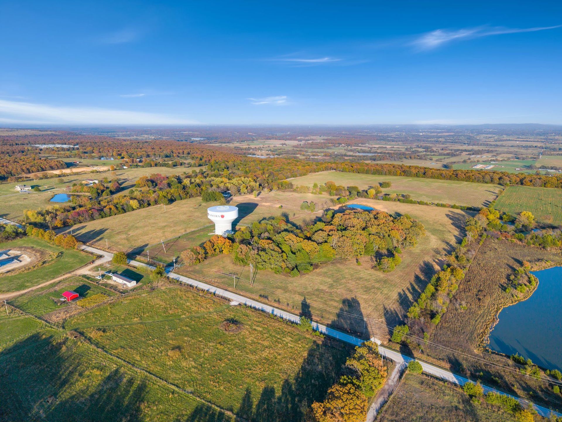 Aerial view of rural landscape with water tower, fields, ponds, and fall foliage under a clear blue sky.