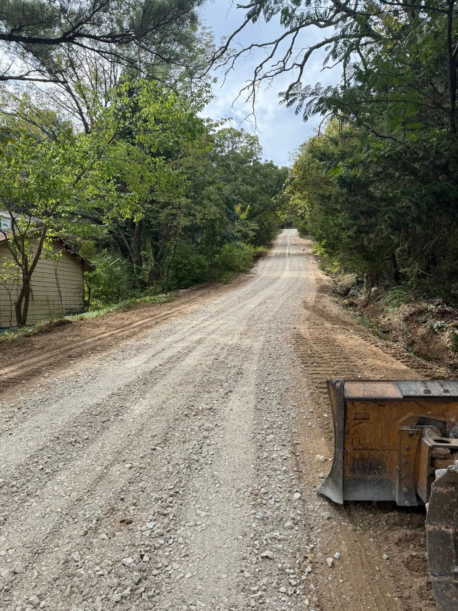 Gravel road through a wooded area.  A piece of construction equipment is on the right side of the road.