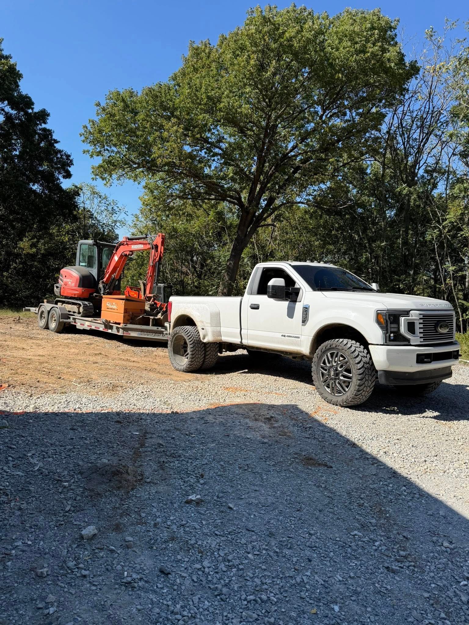 White pickup truck towing a small excavator on a trailer on a gravel driveway, trees in the background.