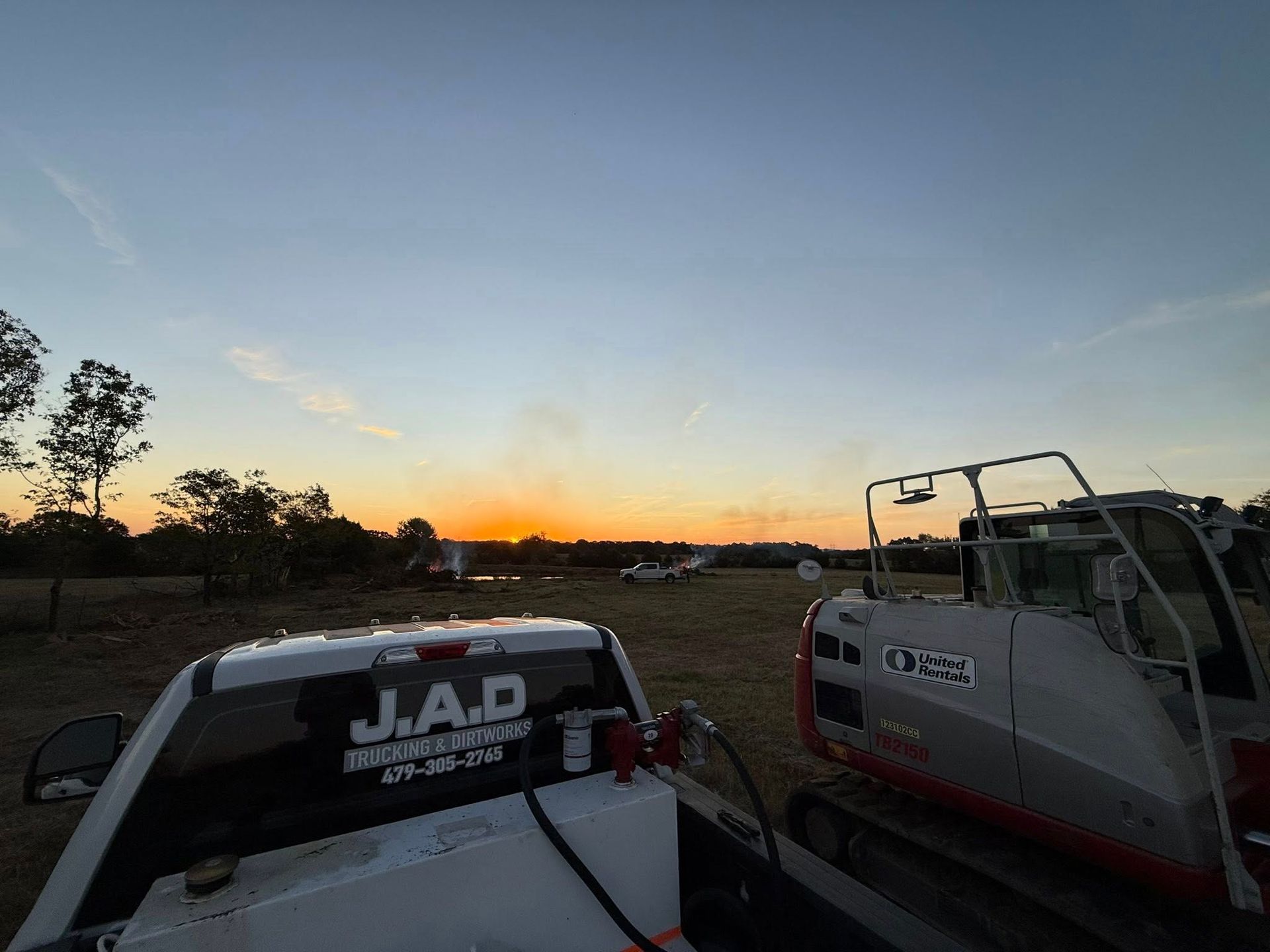 Truck and excavator in field at sunset, J.A.D. logo on truck.