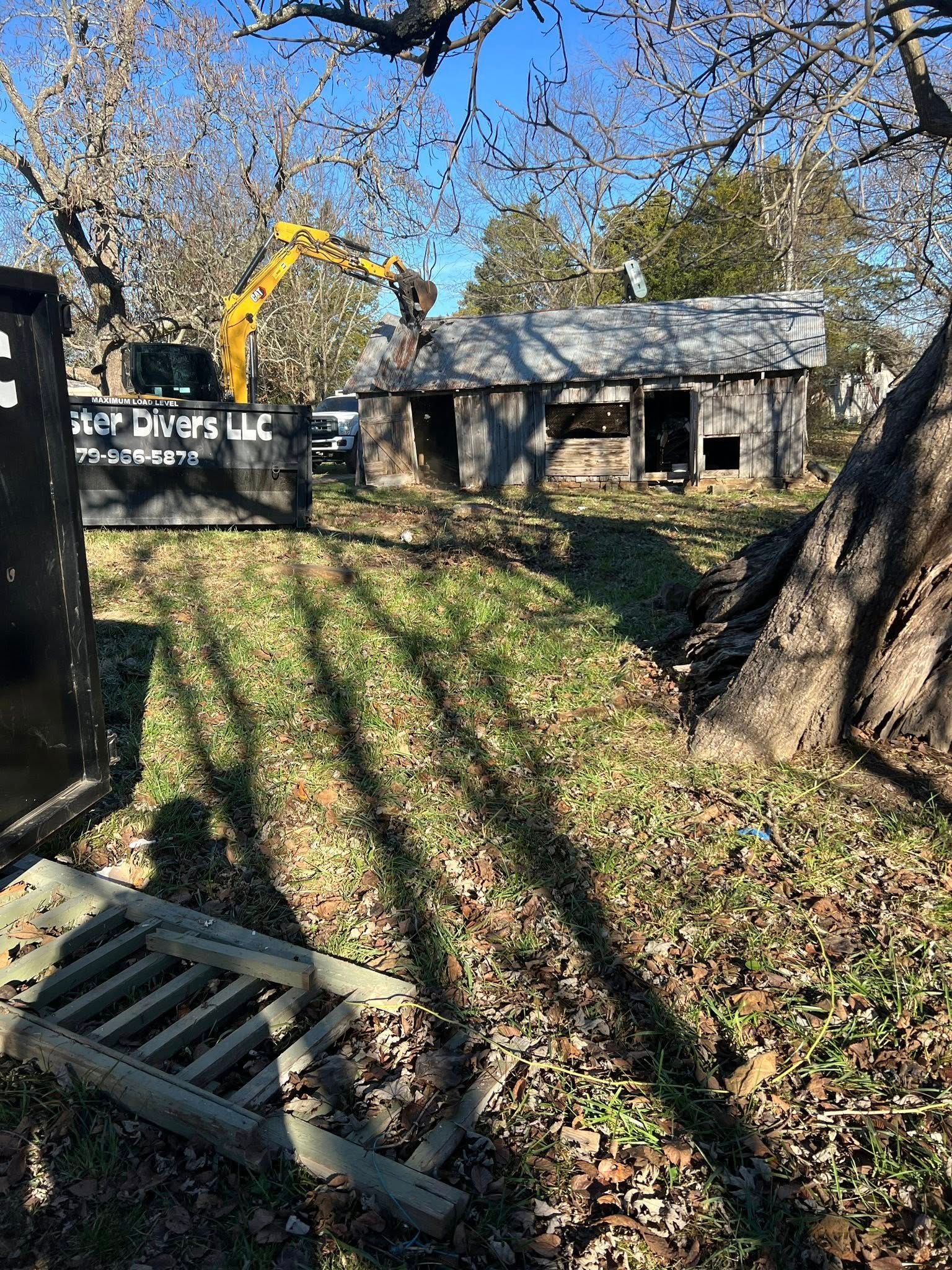 An old, weathered wooden shed being demolished by a yellow excavator on a sunny day.