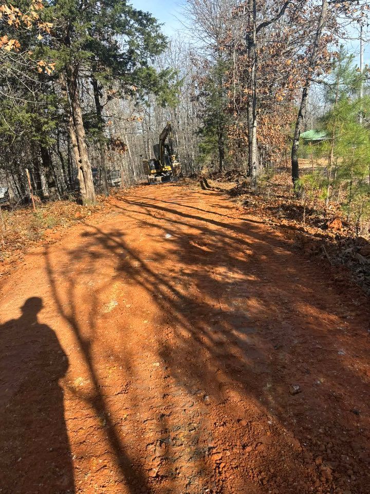 Red dirt path through a forest, shadows cast by trees and a person on a sunny day.