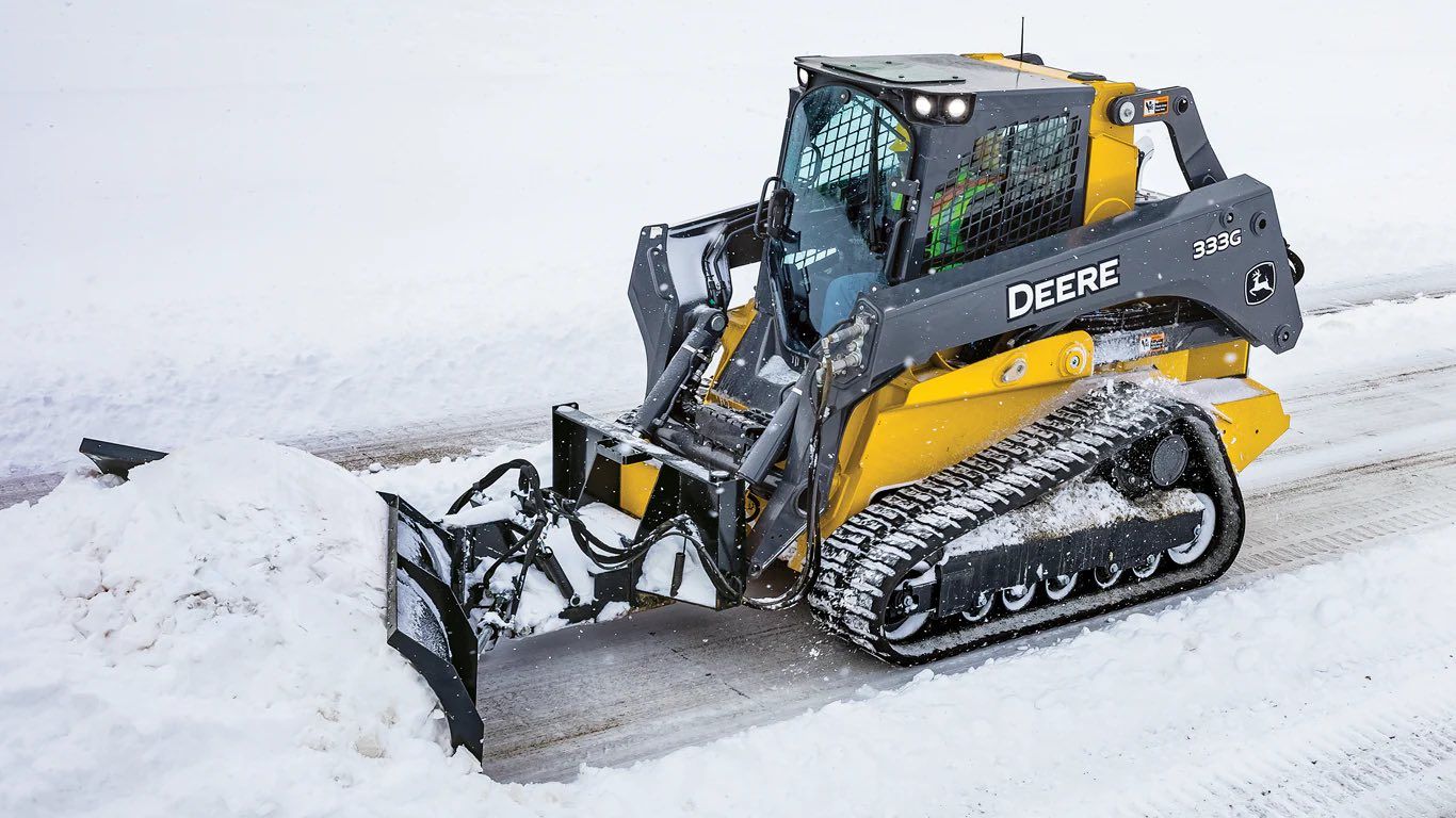 Yellow John Deere skid steer with snow blower clearing snow from a road.