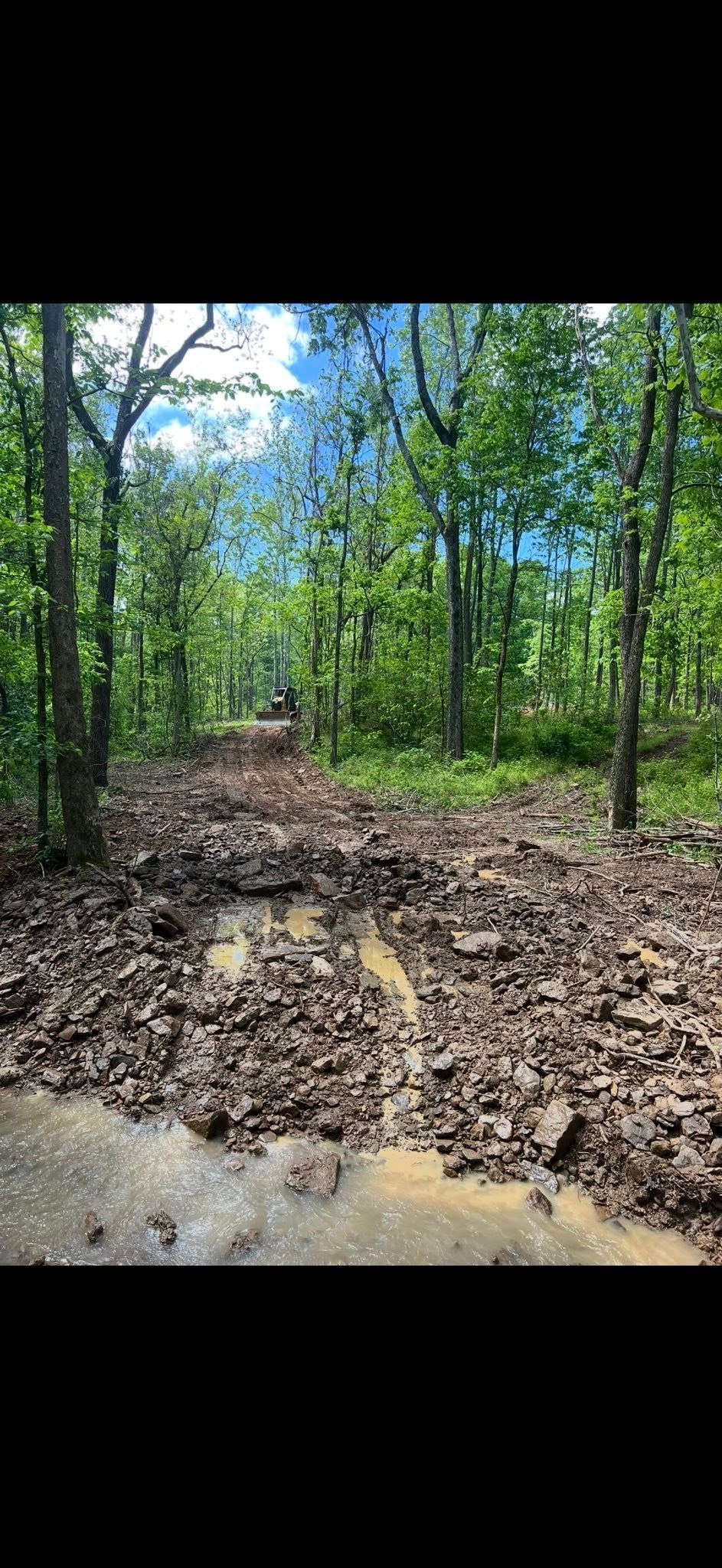 A muddy forest trail with large rocks and trees on both sides. Bright sky visible at the end of the trail.