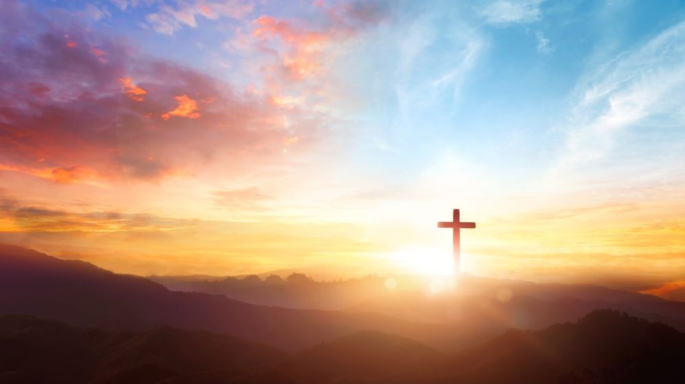 A cross on top of a mountain at sunset with the sun shining through the clouds.