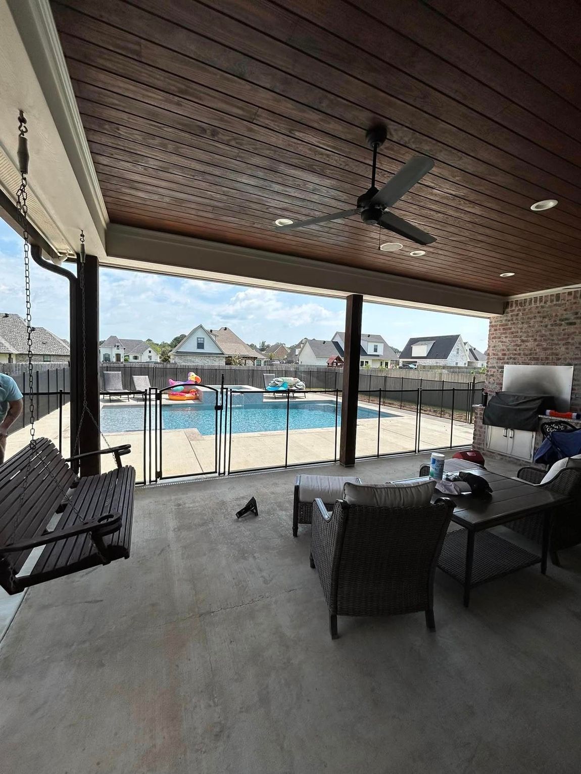 A patio with a swing , chairs , and a ceiling fan overlooking a pool.