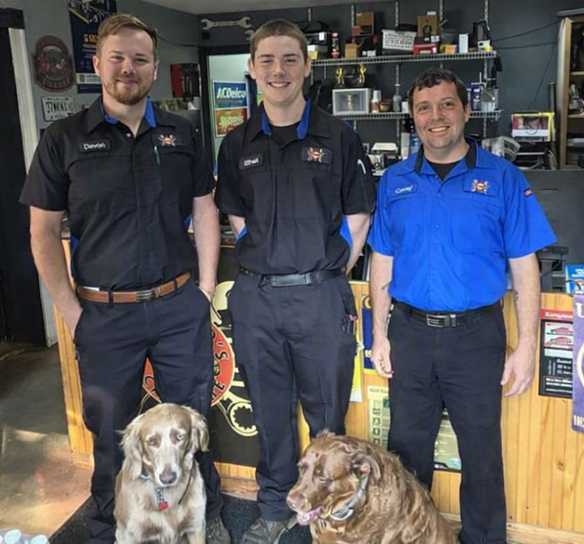 Three men in black and blue shirts stand behind two dogs inside a shop or bar. | Camirands Car Care