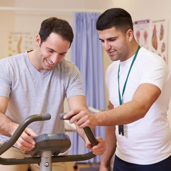 NDIS capacity building. A male trainer helps another man on an exercise bike.
