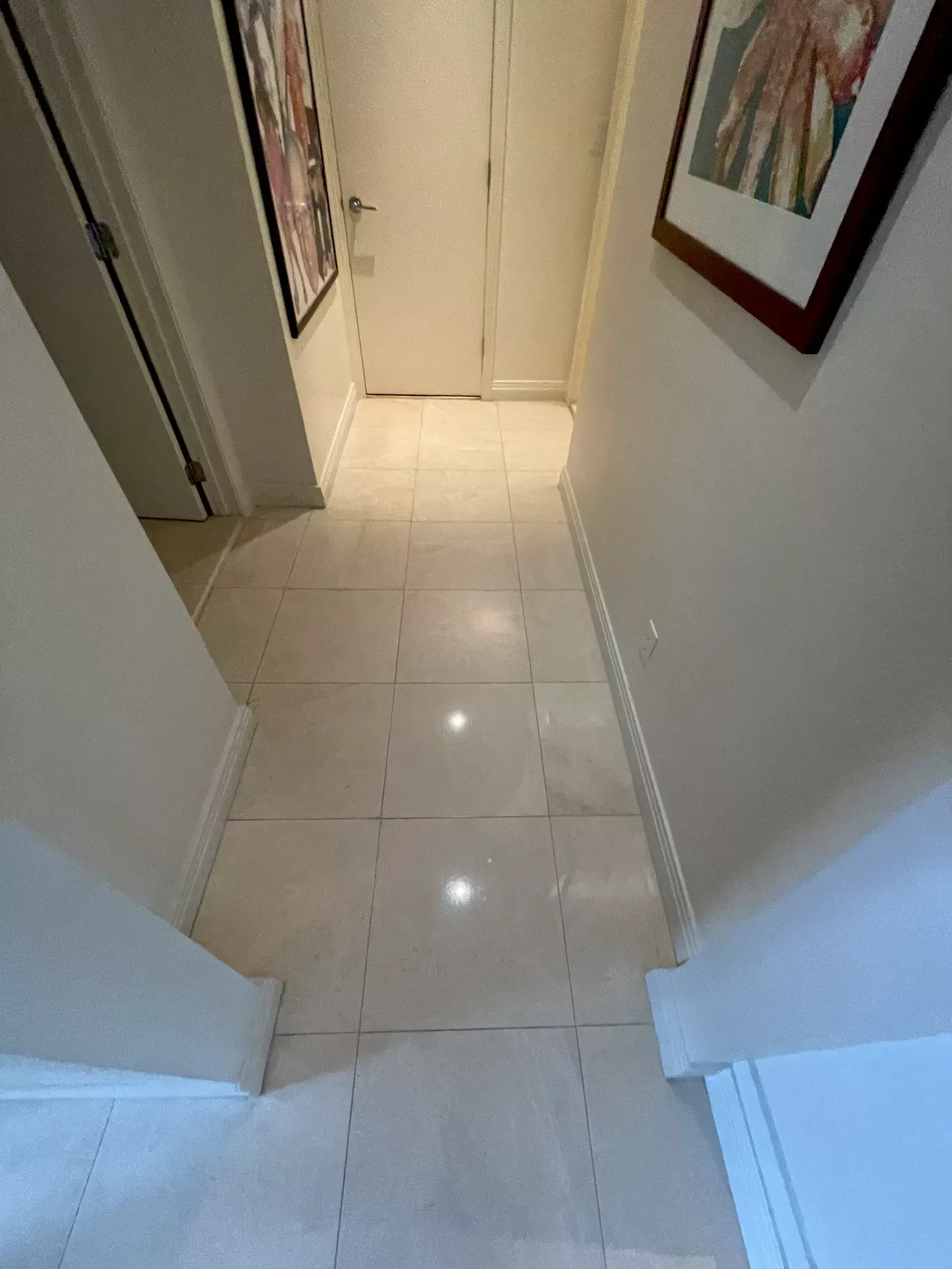Narrow white-tiled hallway with a door at the end and a framed picture on the wall.