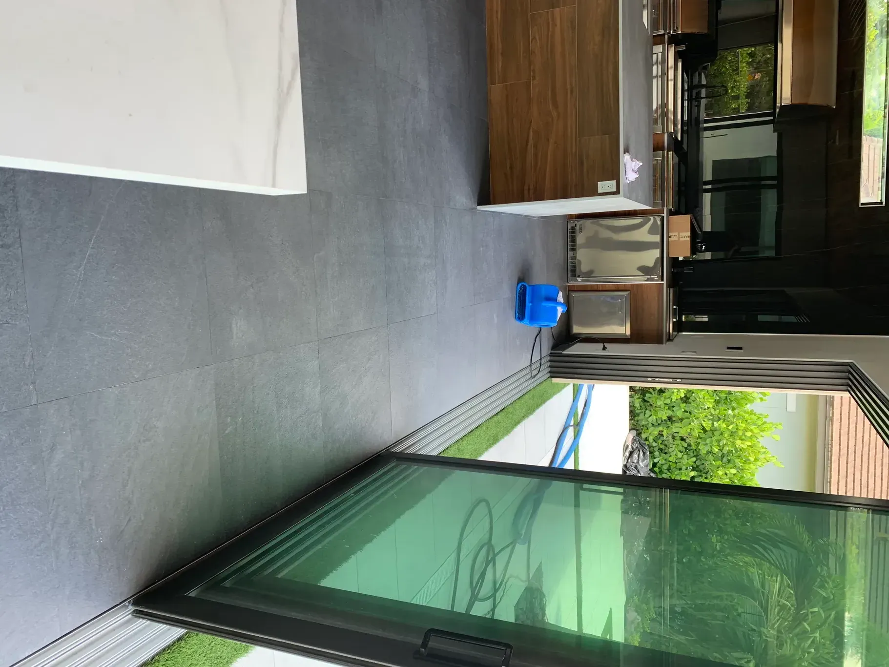 Covered outdoor patio with gray floor, green glass wall, blue bucket, and stacked boxes near a doorway