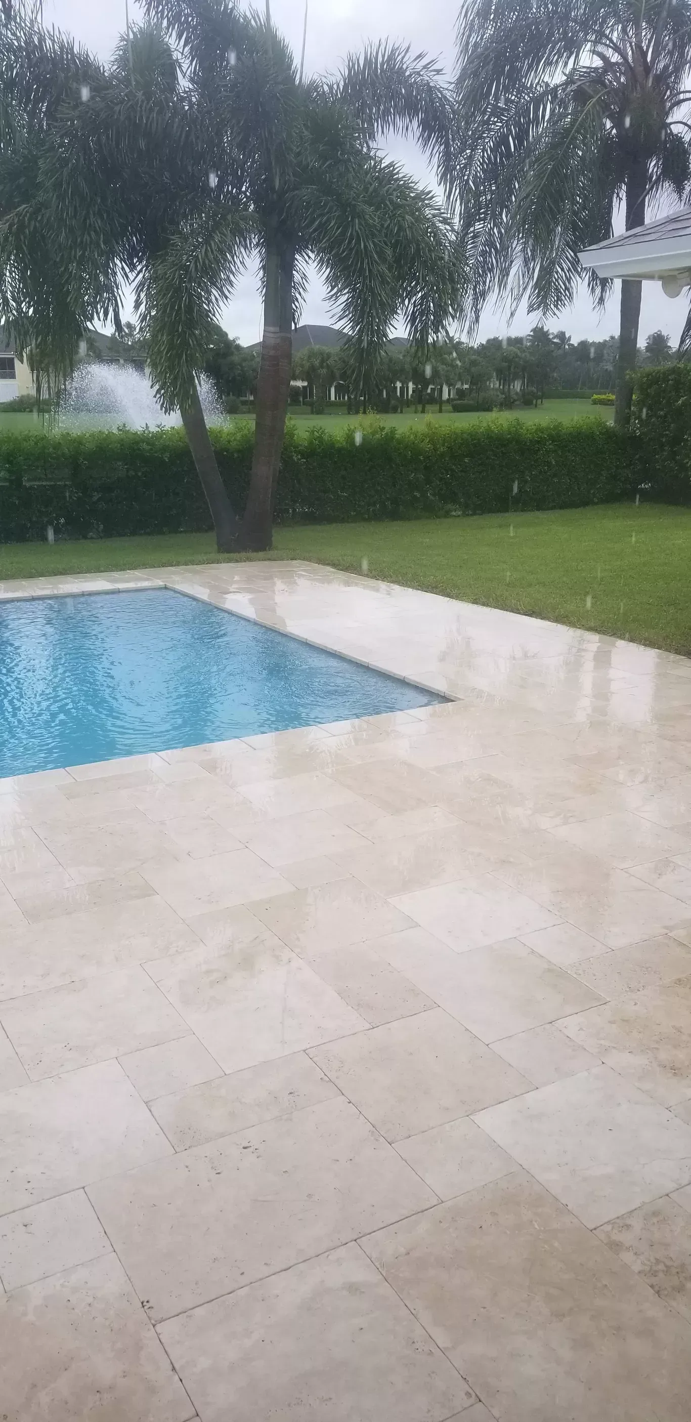 Rainy poolside patio with beige stone tiles, blue pool, and green lawn and trees in the background
