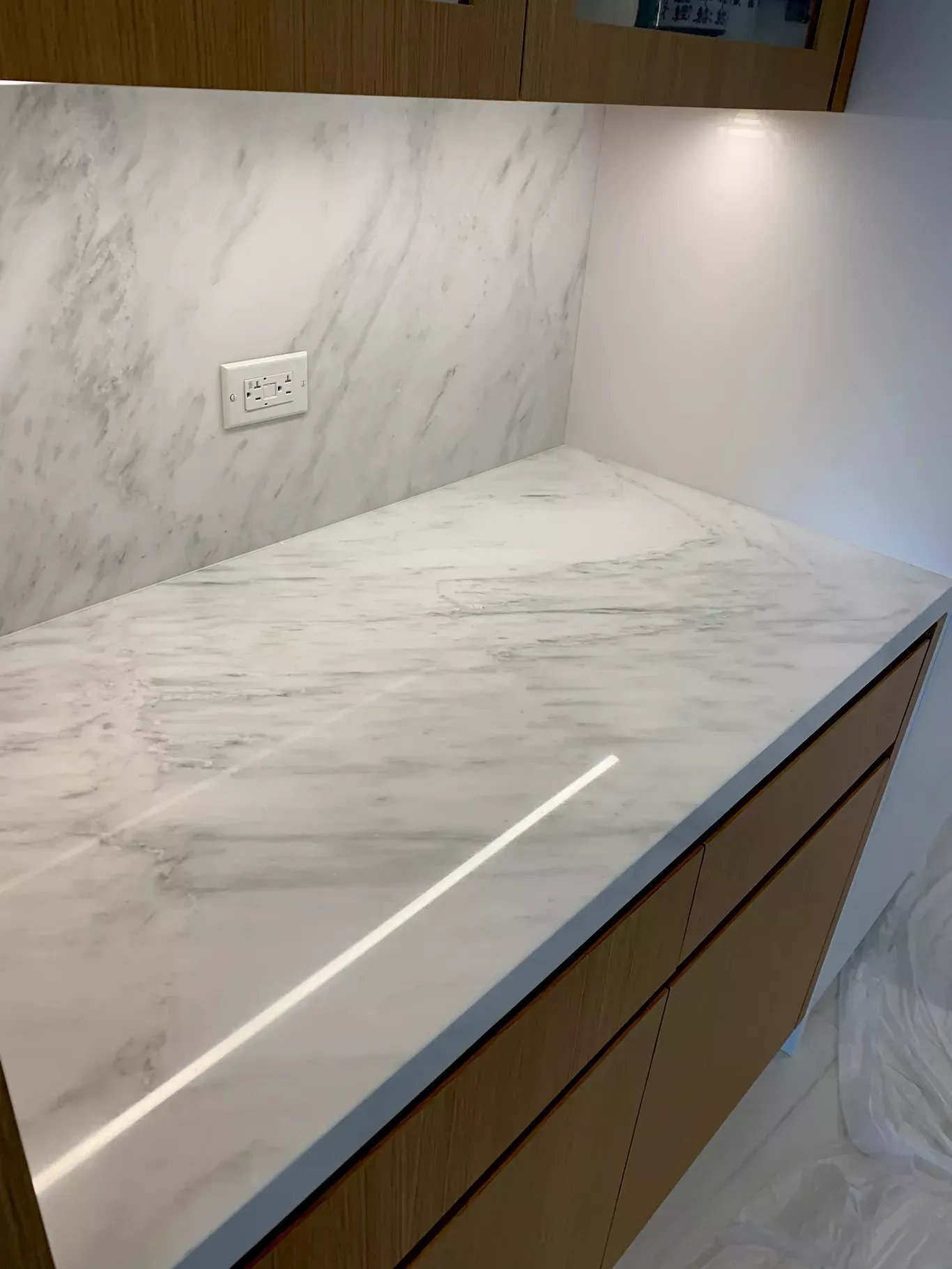 Marble-look kitchen countertop with wood cabinets, backsplash, and a wall outlet under bright lighting
