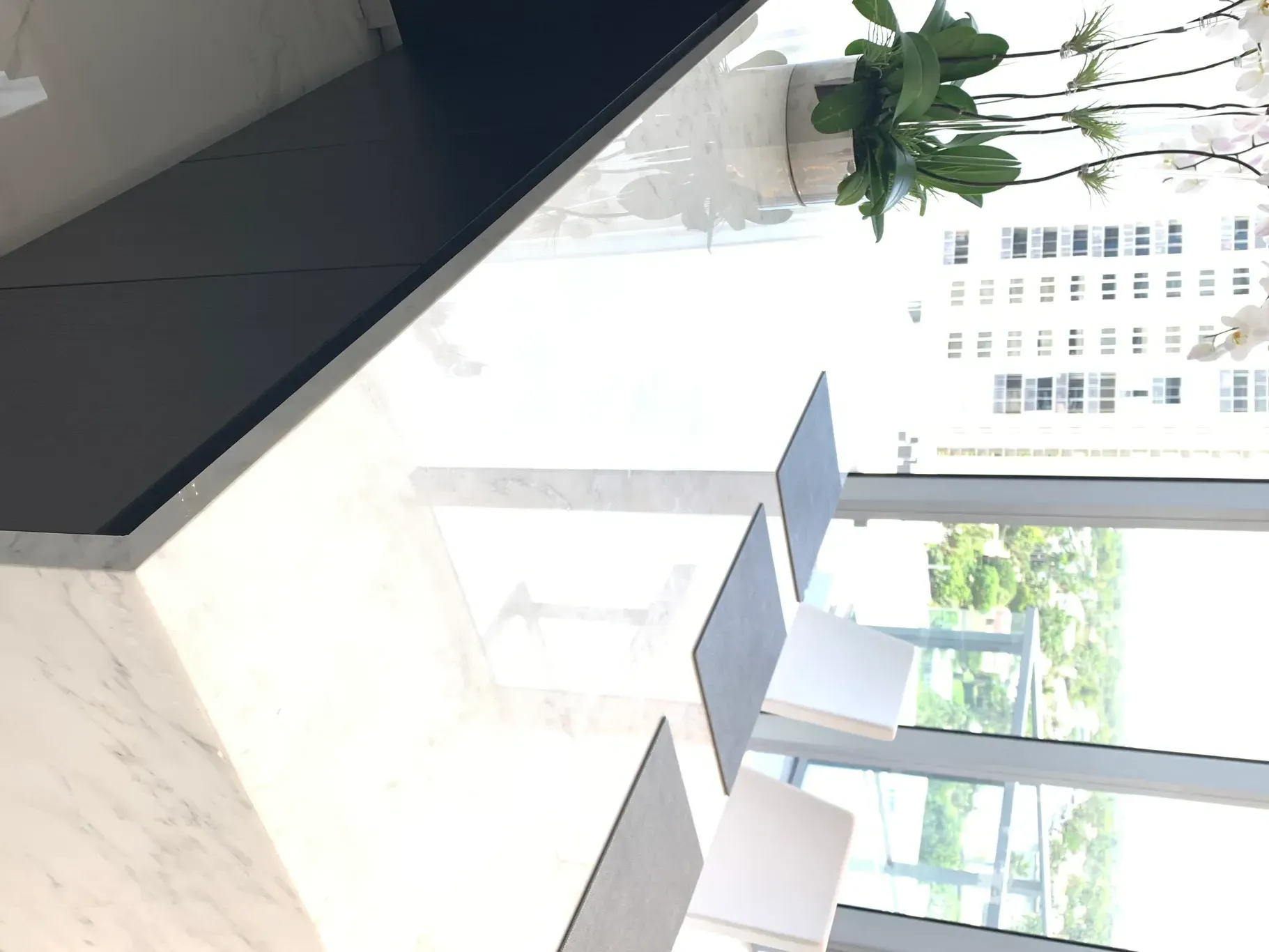 Modern kitchen with black island, white chairs, marble counters, and large windows overlooking greenery