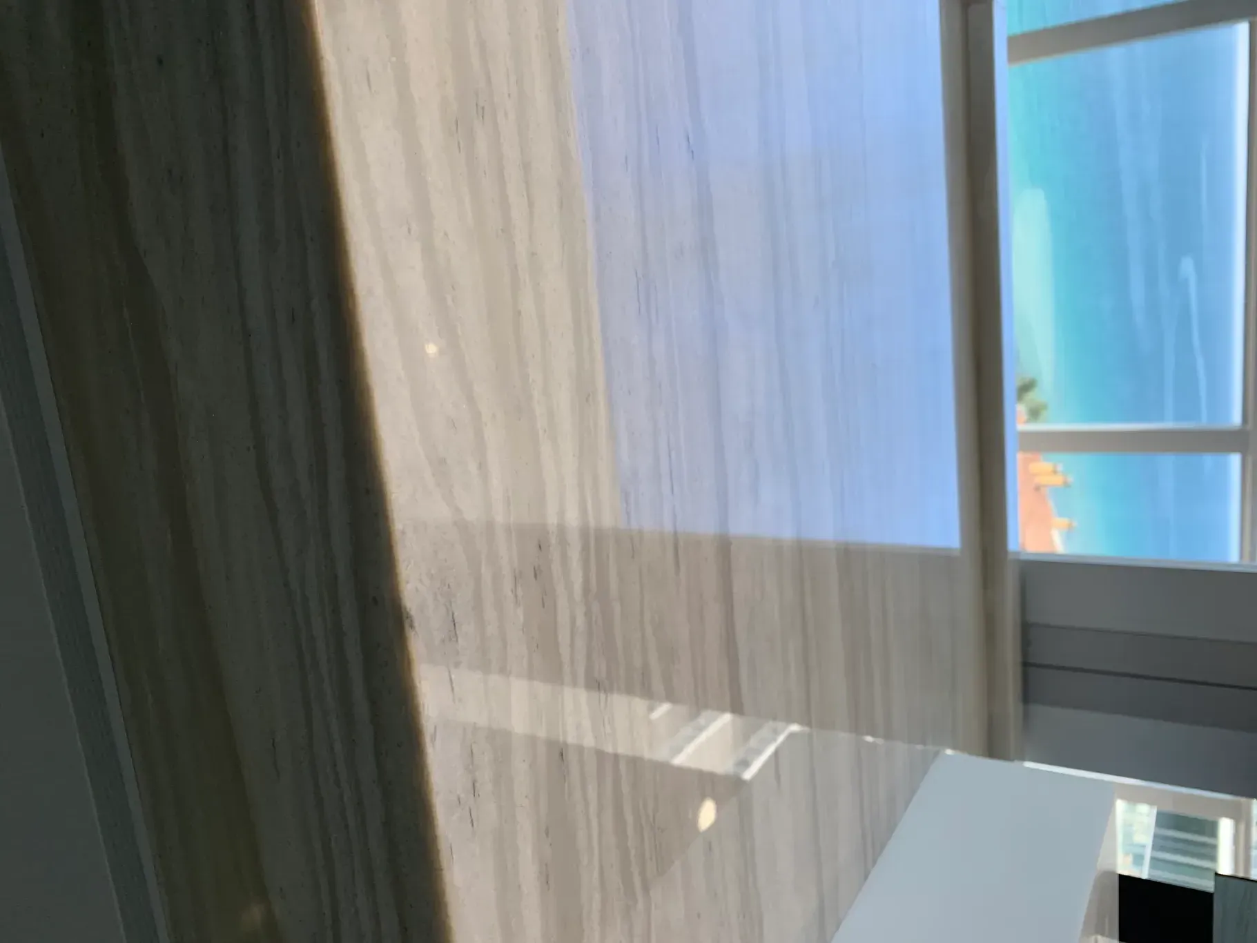 Sunlit room with curtains beside a window and a white desk, overlooking blue water outside