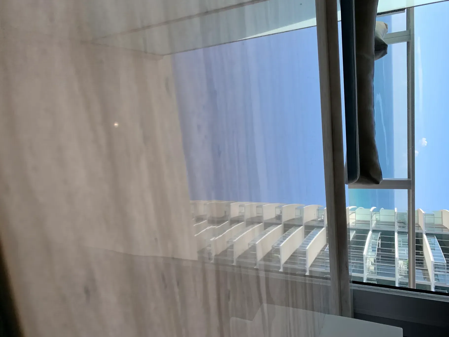 Interior view through a sheer curtain toward a balcony, with blue sky and buildings outside.