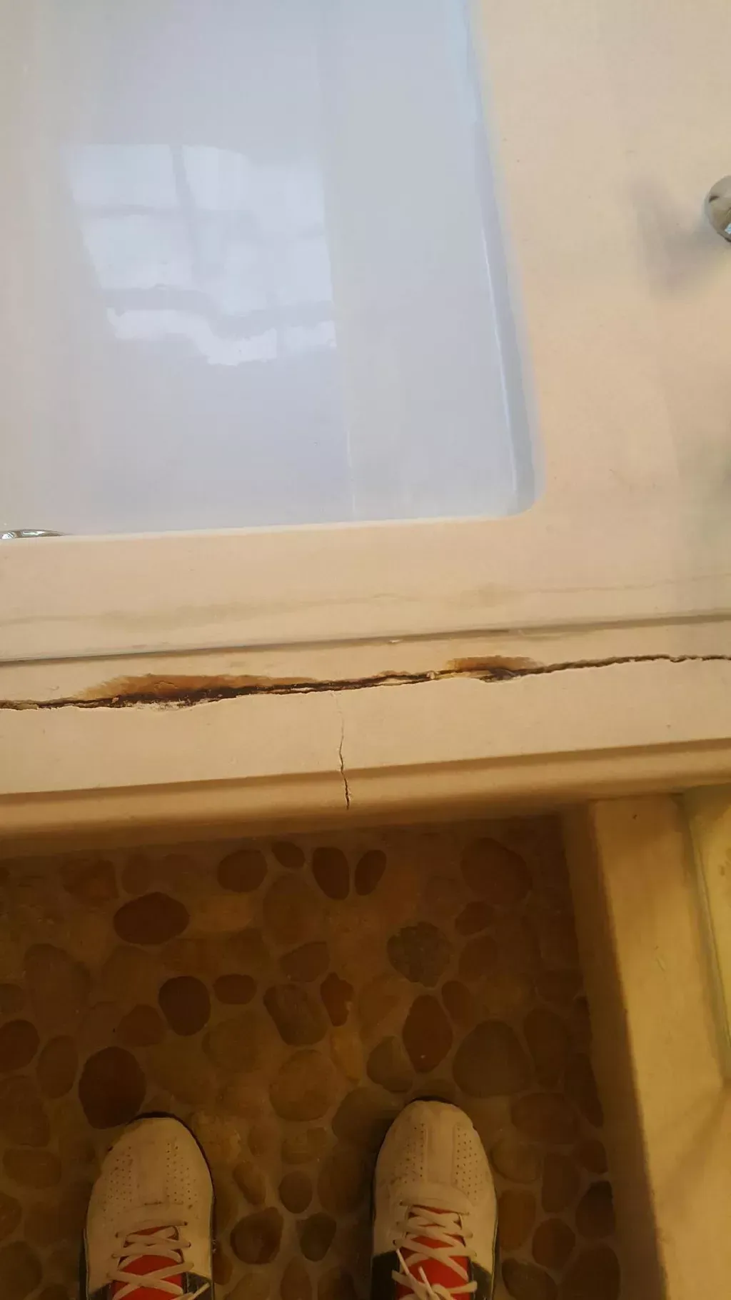 White cabinet door with a long crack near the bottom, viewed above patterned floor and white shoes.