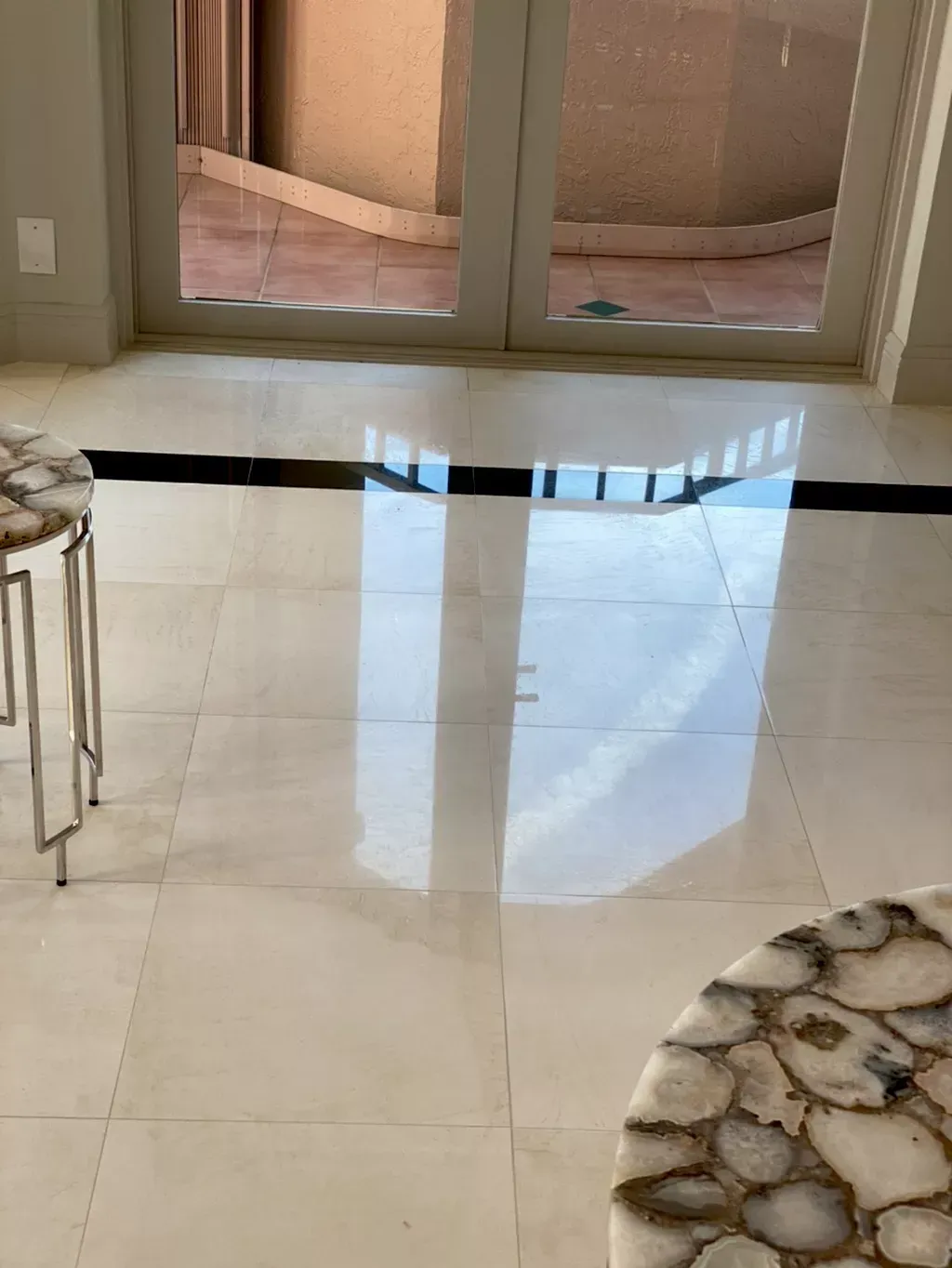 Shiny beige tiled floor with reflections near a window and a small stool at left