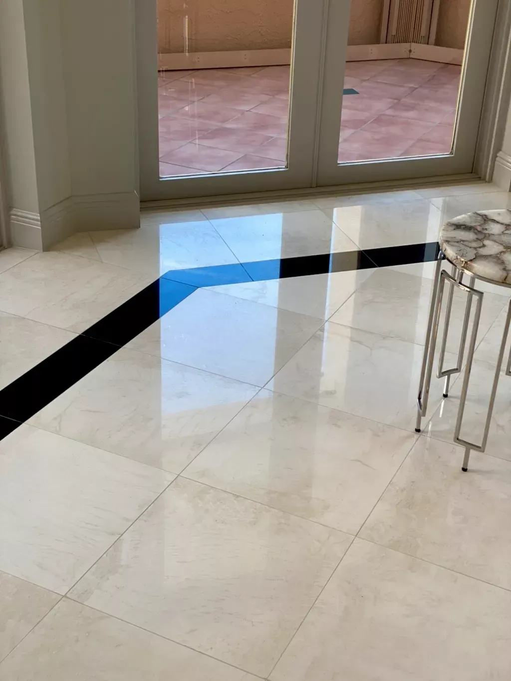 Glossy tiled floor with black border near glass doors and a small metal side table