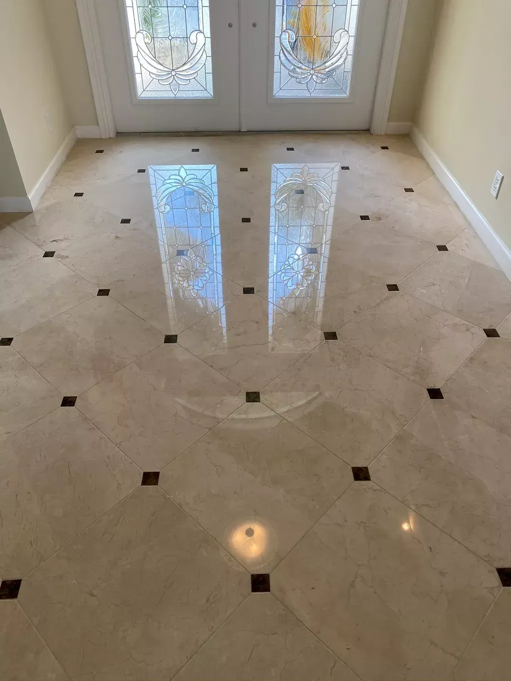 Polished beige tiled floor with small dark accents and reflected light near double glass doors.