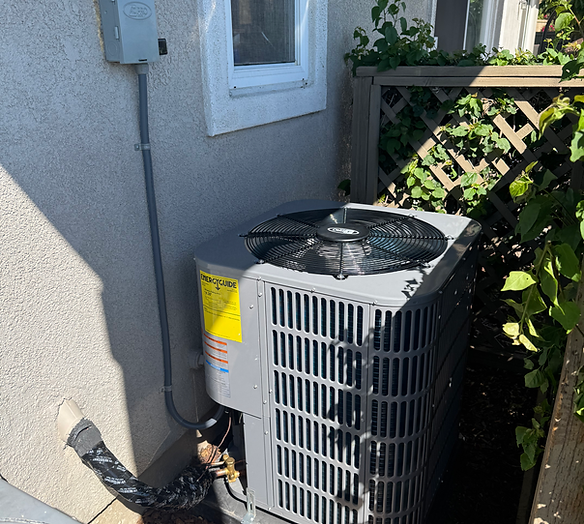 An outdoor air conditioning unit sits against the side of a house, connected by conduit to a wall-mounted electrical box.
