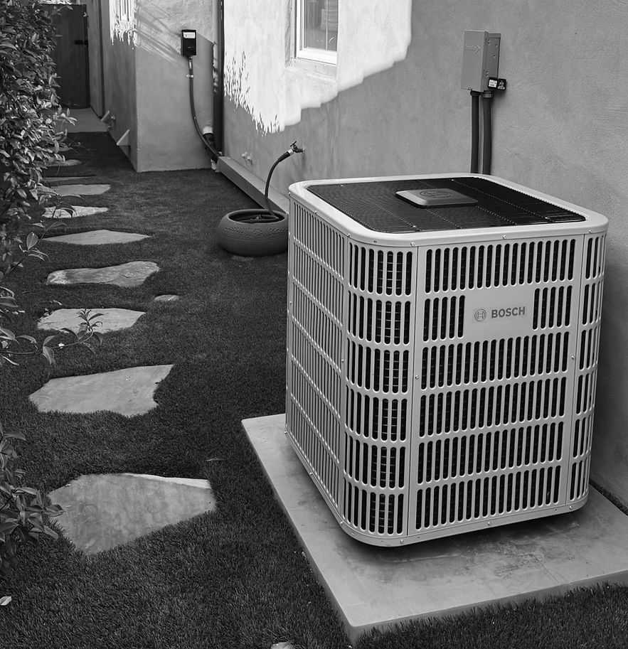 A Bosch air conditioner unit sits on a concrete pad next to a wall, adjacent to a stone-path lawn.