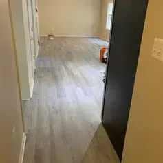 Hallway with light gray wood-look flooring, leading to a room with beige walls and a window. Black cabinet on the right.