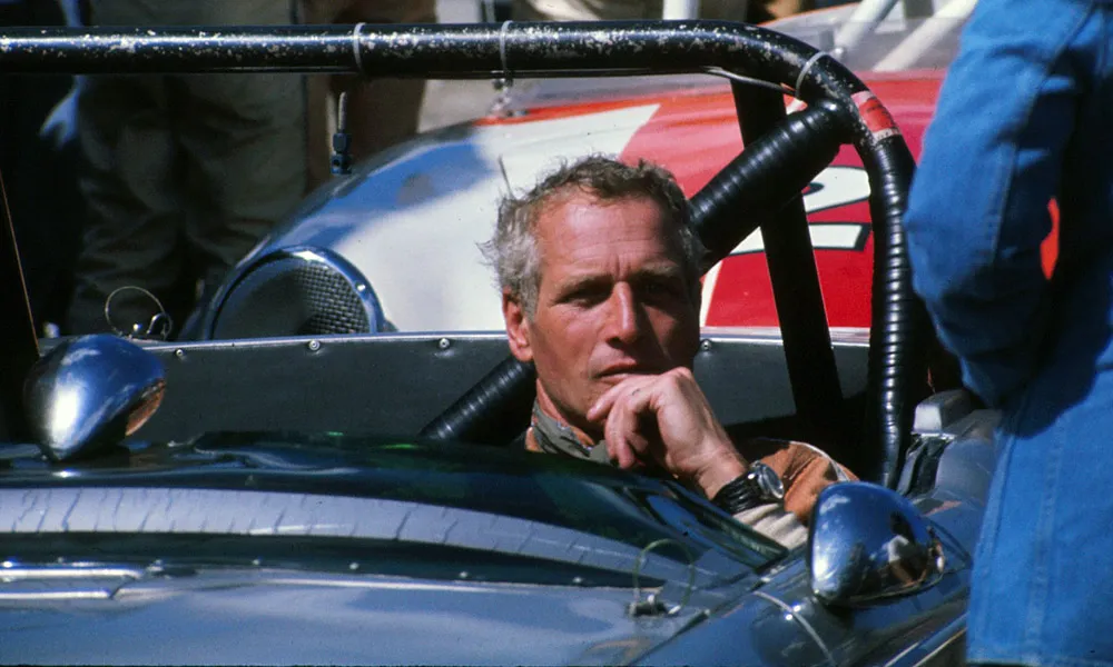 Paul Newman in a race car, resting his chin on his hand, at the track.