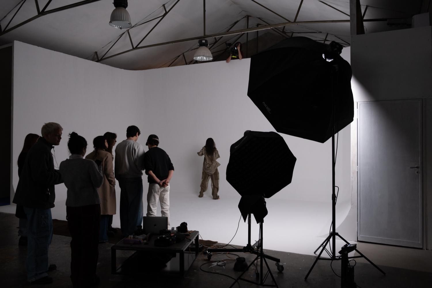 Una troupe cinematografica osserva una persona in posa in uno studio luminoso e bianco, attrezzato con grandi ombrelli per l'illuminazione.