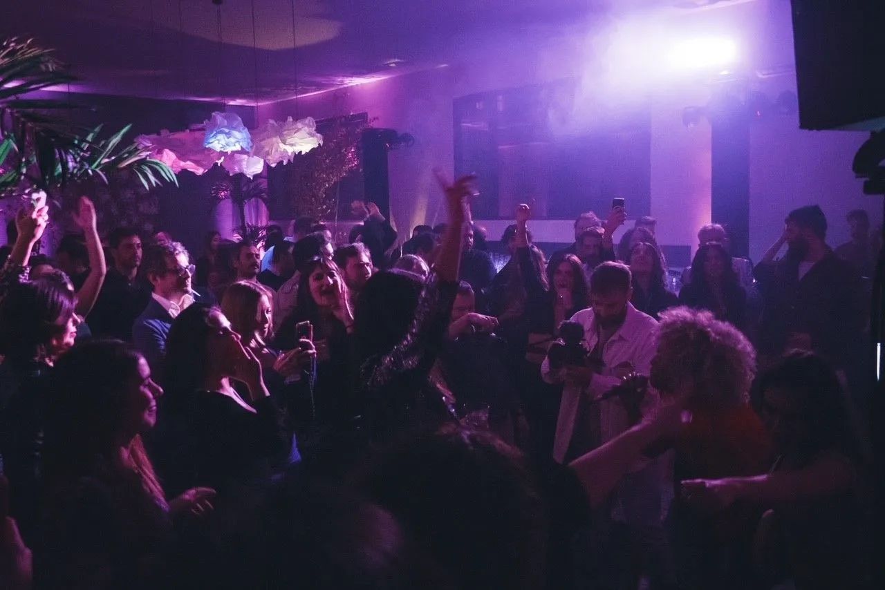 Una discoteca affollata con luci soffuse viola, gente che balla, alza le mani e socializza.