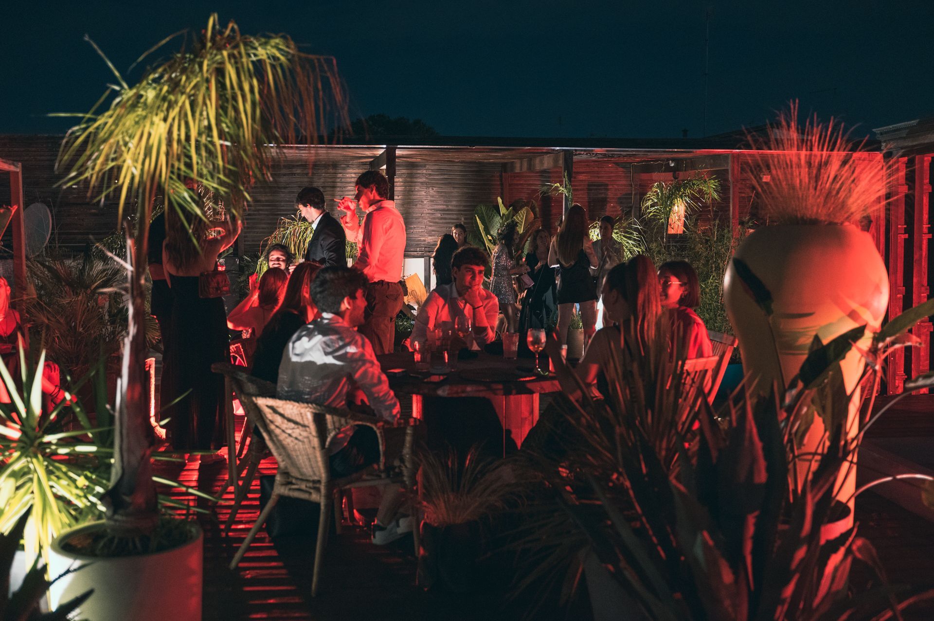 Di notte, le persone si riuniscono su una terrazza all'aperto buia e ricca di piante, illuminata da una luce calda e soffusa.