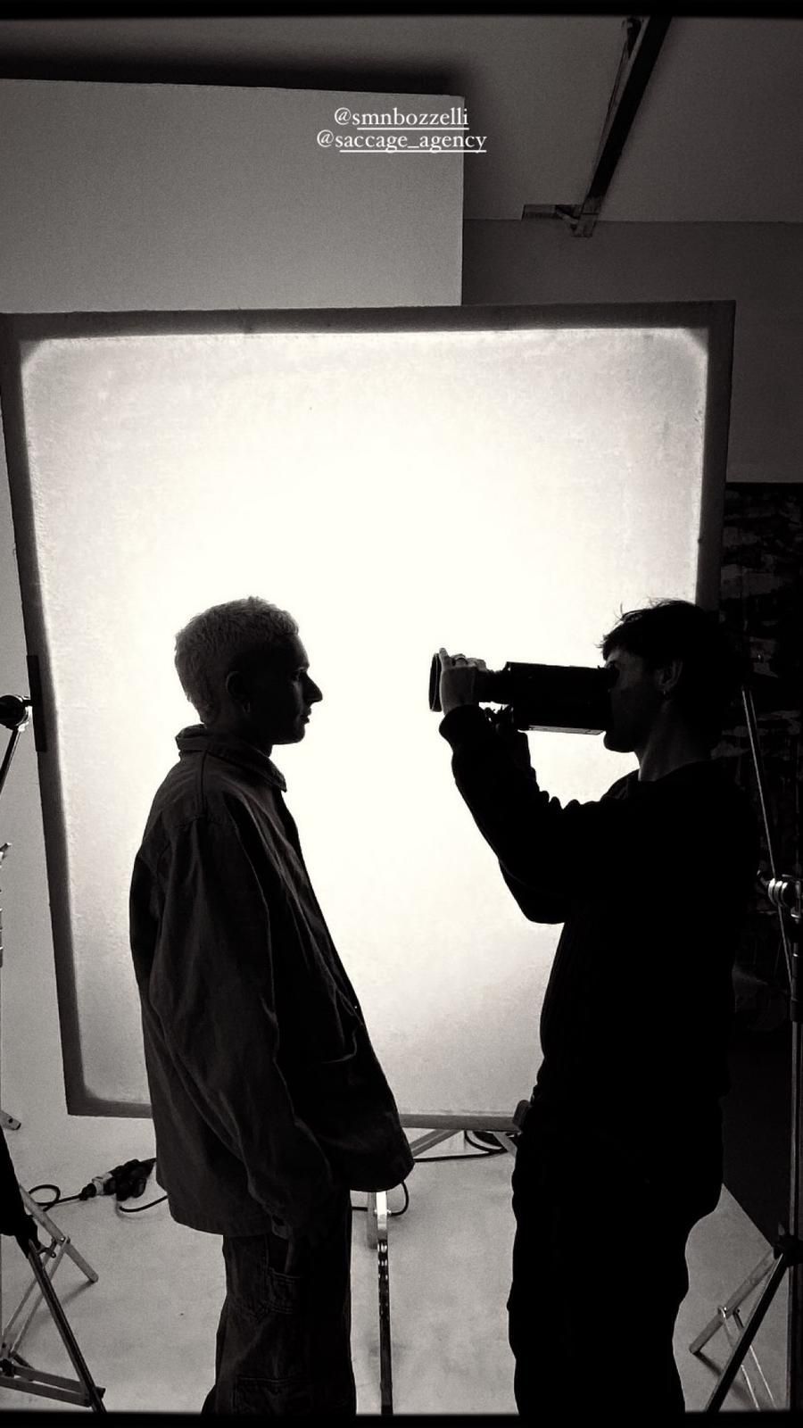 Una troupe cinematografica riprende un gruppo eterogeneo di sei persone sedute su un grande divano in un soggiorno interno ben illuminato.