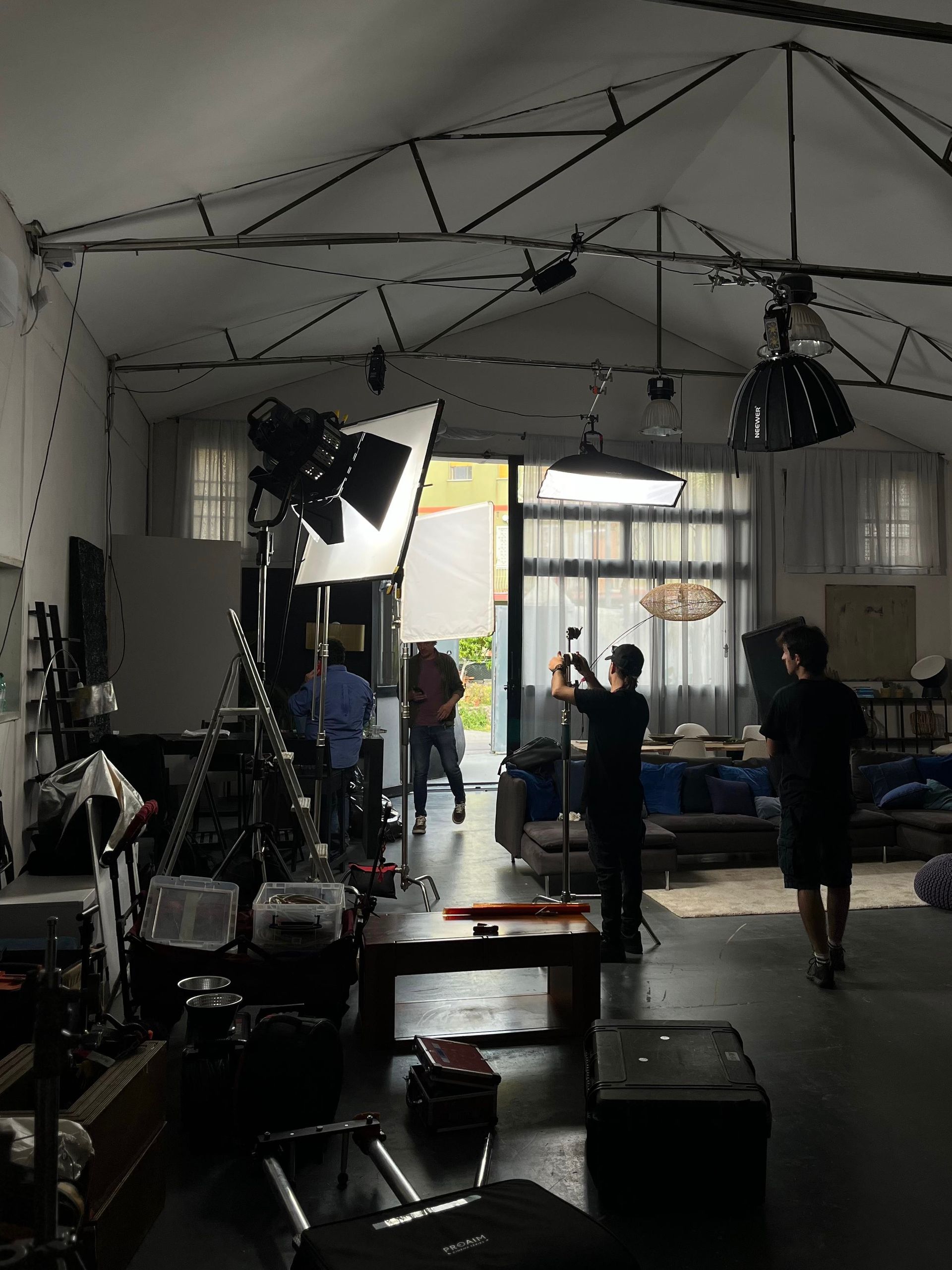 Un membro della troupe cinematografica manovra una telecamera su un treppiede, riprendendo quattro persone sedute su un divano in uno studio ben illuminato.