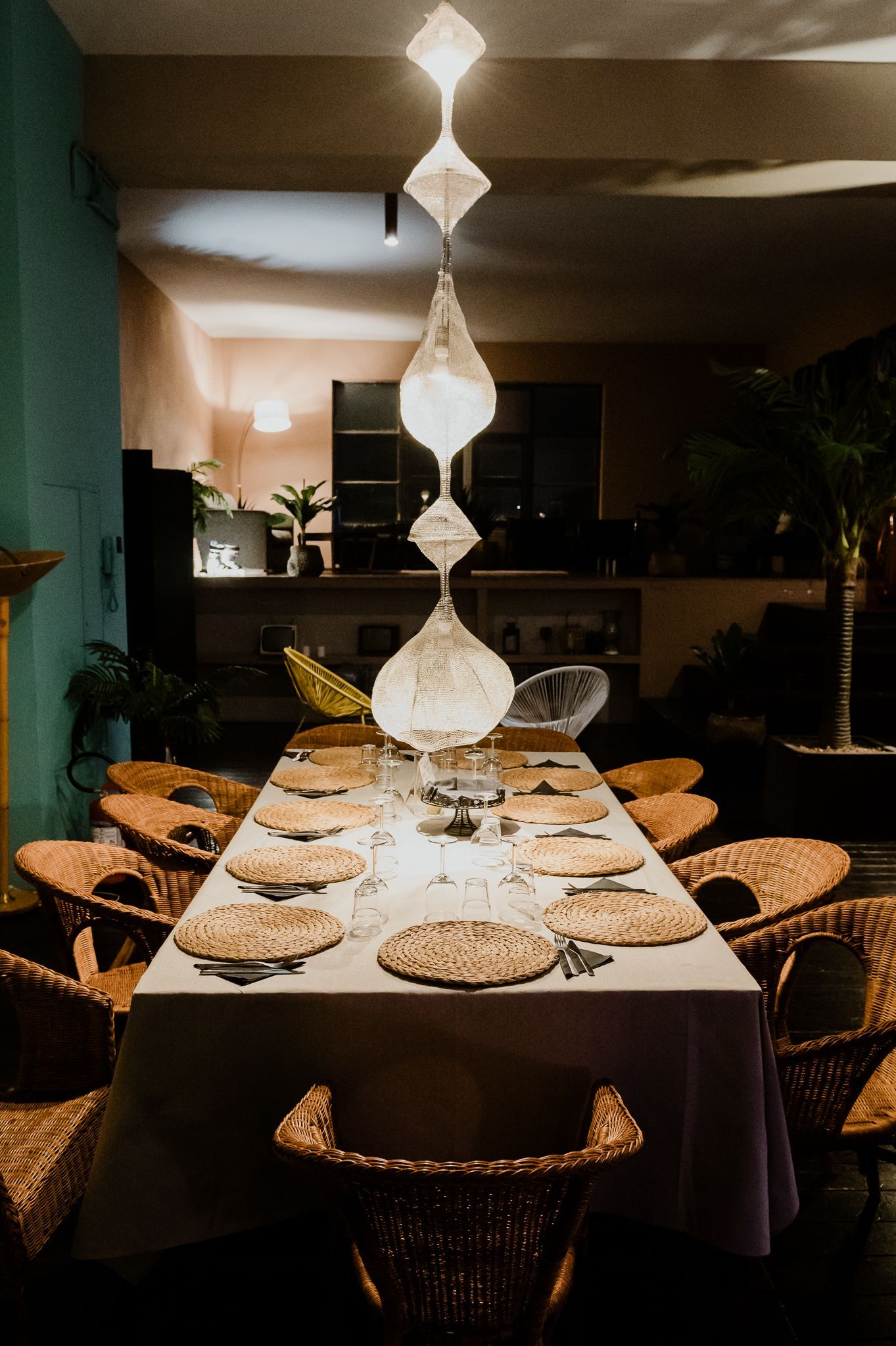 Un tavolo da pranzo con sedie intrecciate è posizionato sotto lampade a sospensione, contro una parete verde tenue decorata con opere d'arte astratte.
