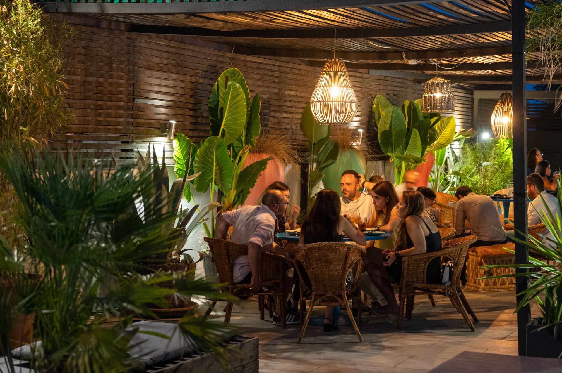 Un gruppo di persone cena in un ristorante ben illuminato con pavimenti in legno scuro, sedie rustiche in rattan e illuminazione moderna.
