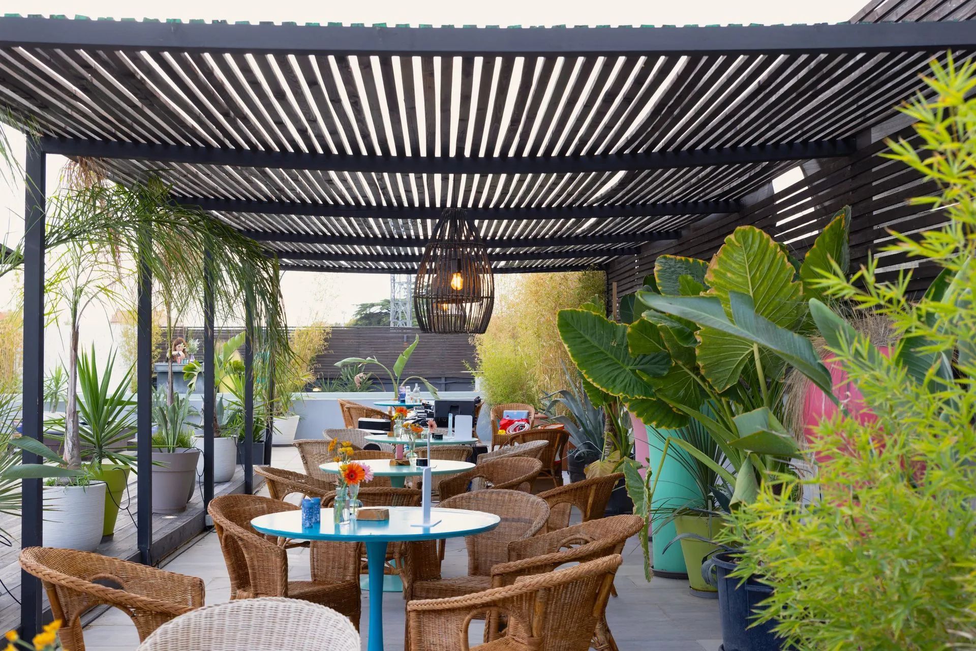 Zona esterna con patio, arredata con sedie in vimini, tavolini blu e rigogliose piante verdi sotto una pergola a doghe nere.