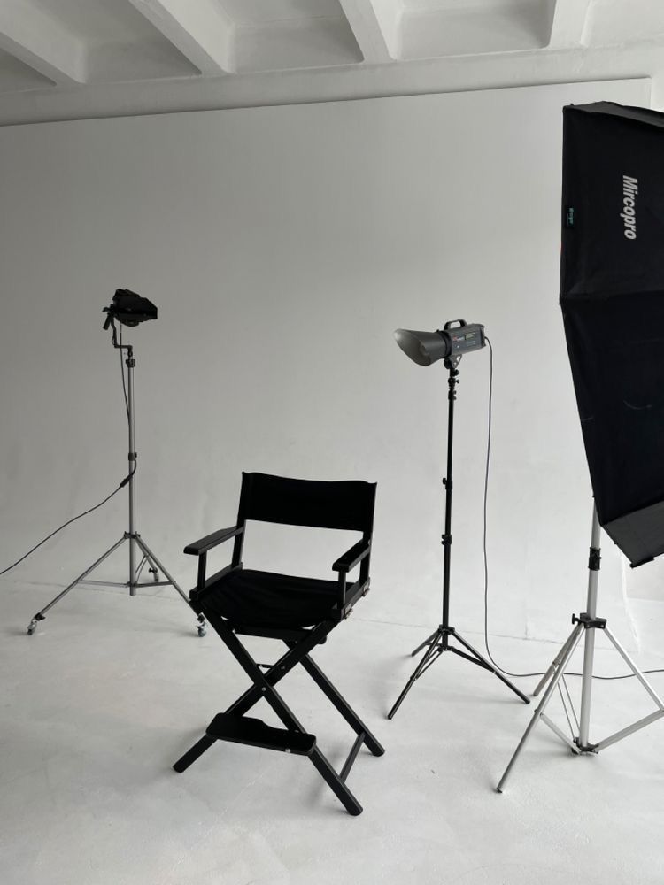 Una troupe cinematografica lavora in uno studio bianco, filmando una persona in piedi davanti a uno sfondo bianco con l'ausilio di attrezzature di illuminazione.