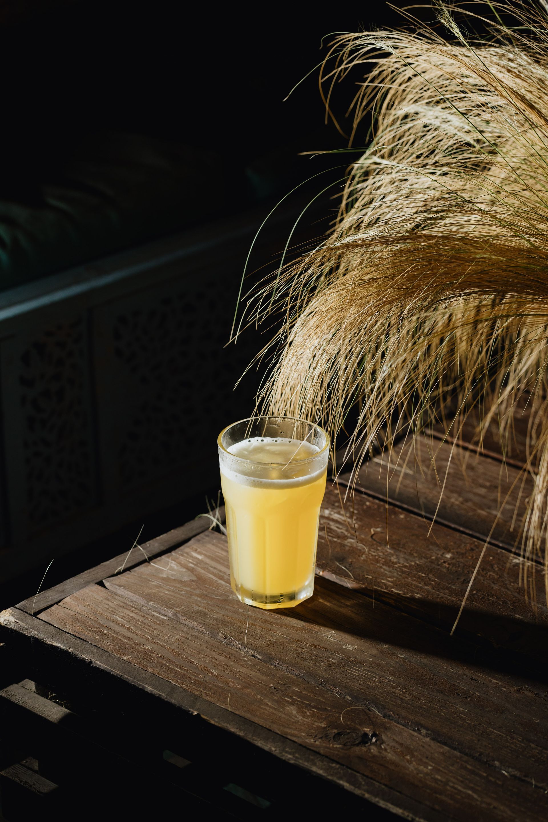 In una stanza scarsamente illuminata, un bicchiere di birra dorata è appoggiato su un rustico tavolo di legno accanto a un mazzo decorativo di spighe di grano essiccate.
