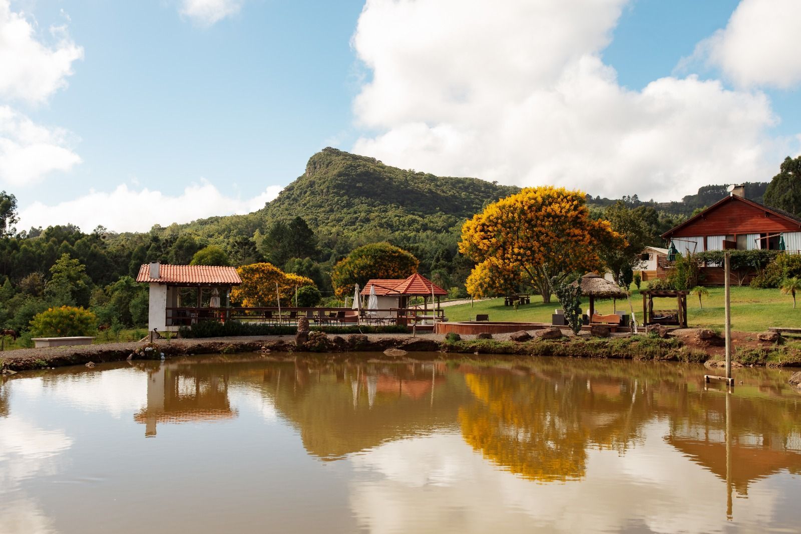 Lagoa da aldeia refletindo árvores, cabanas e uma colina verde sob um céu nublado.