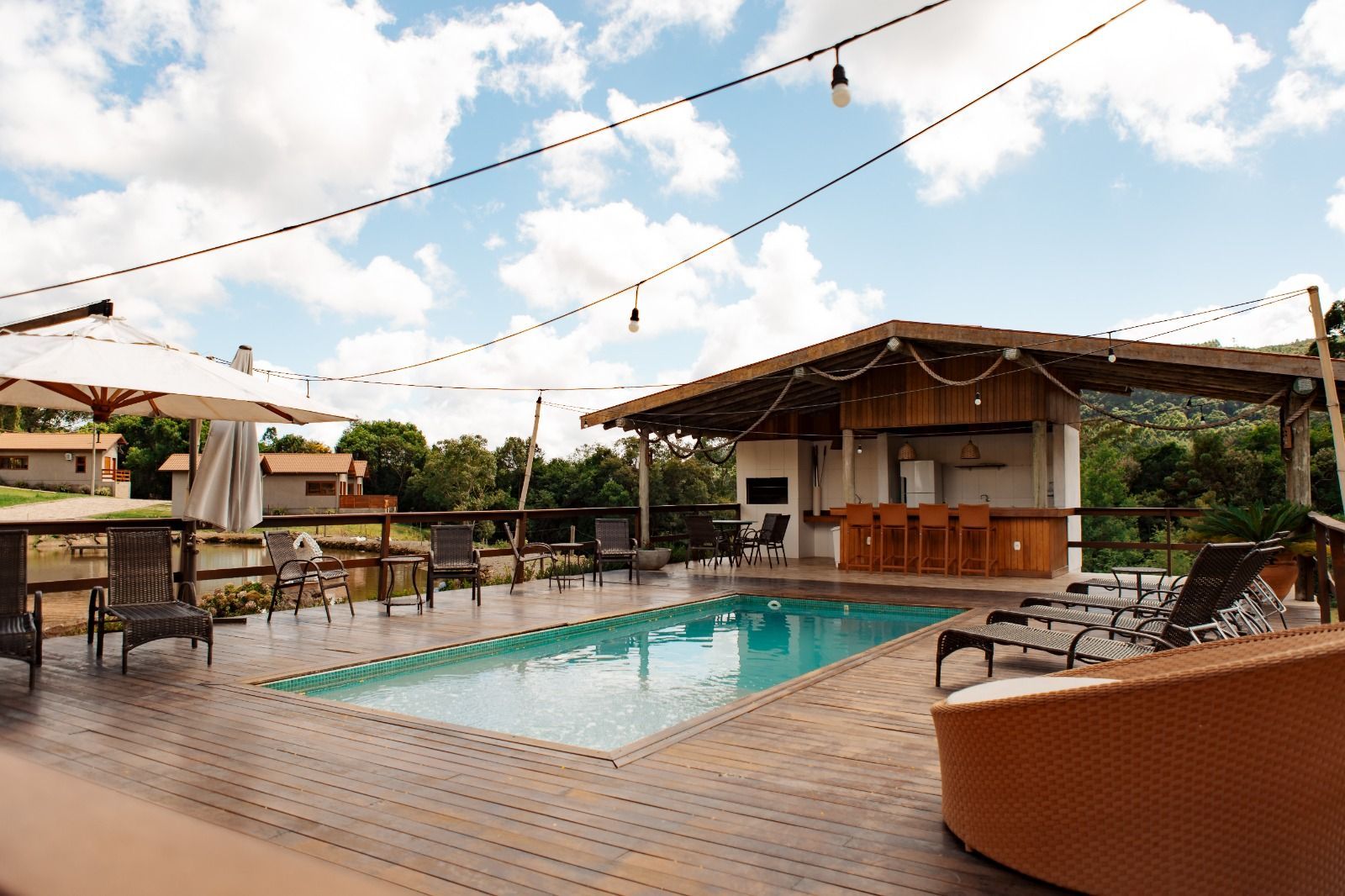 Terraço com piscina na cobertura, espreguiçadeiras, luzes de corda e um pequeno bar sob um céu nublado.