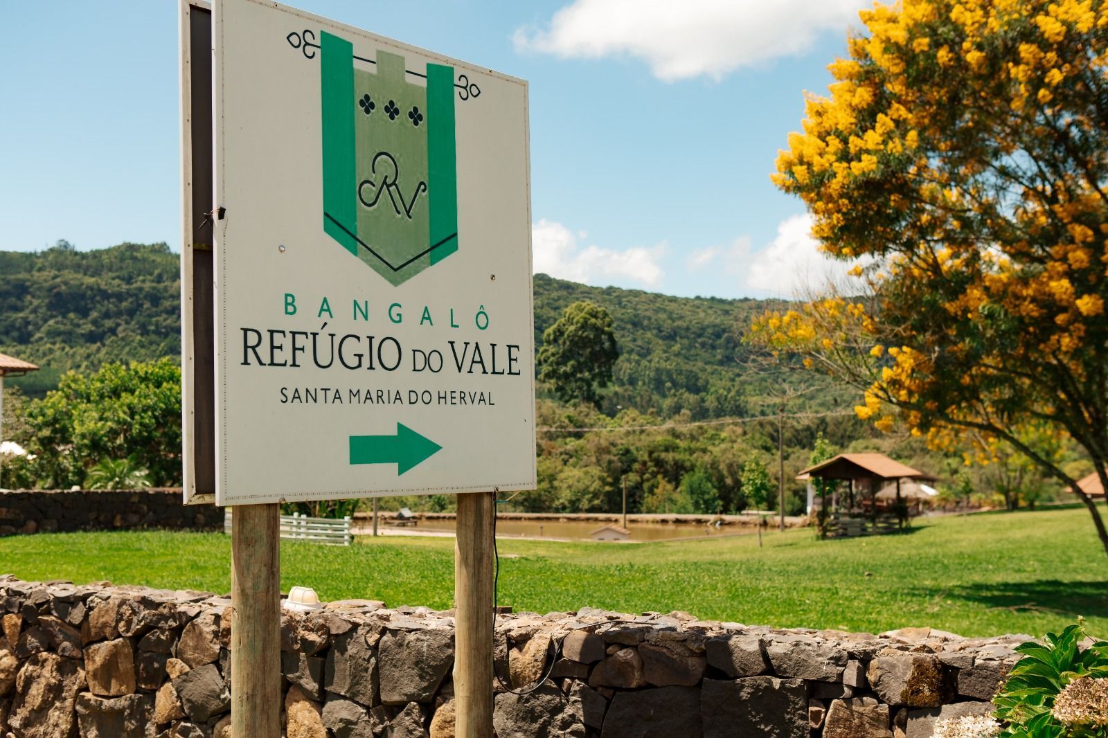 Placa indicativa de Bangalô Refúgio Vale em uma paisagem rural verdejante com árvores e muro de pedra.
