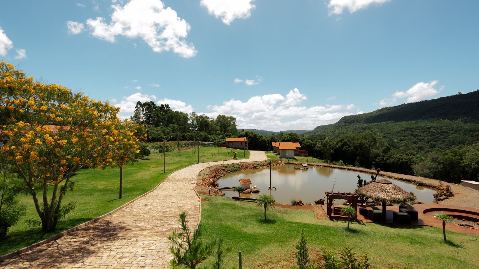 Lagoa rural com caminho de terra, colinas verdejantes e um deck de madeira sob um céu azul brilhante.