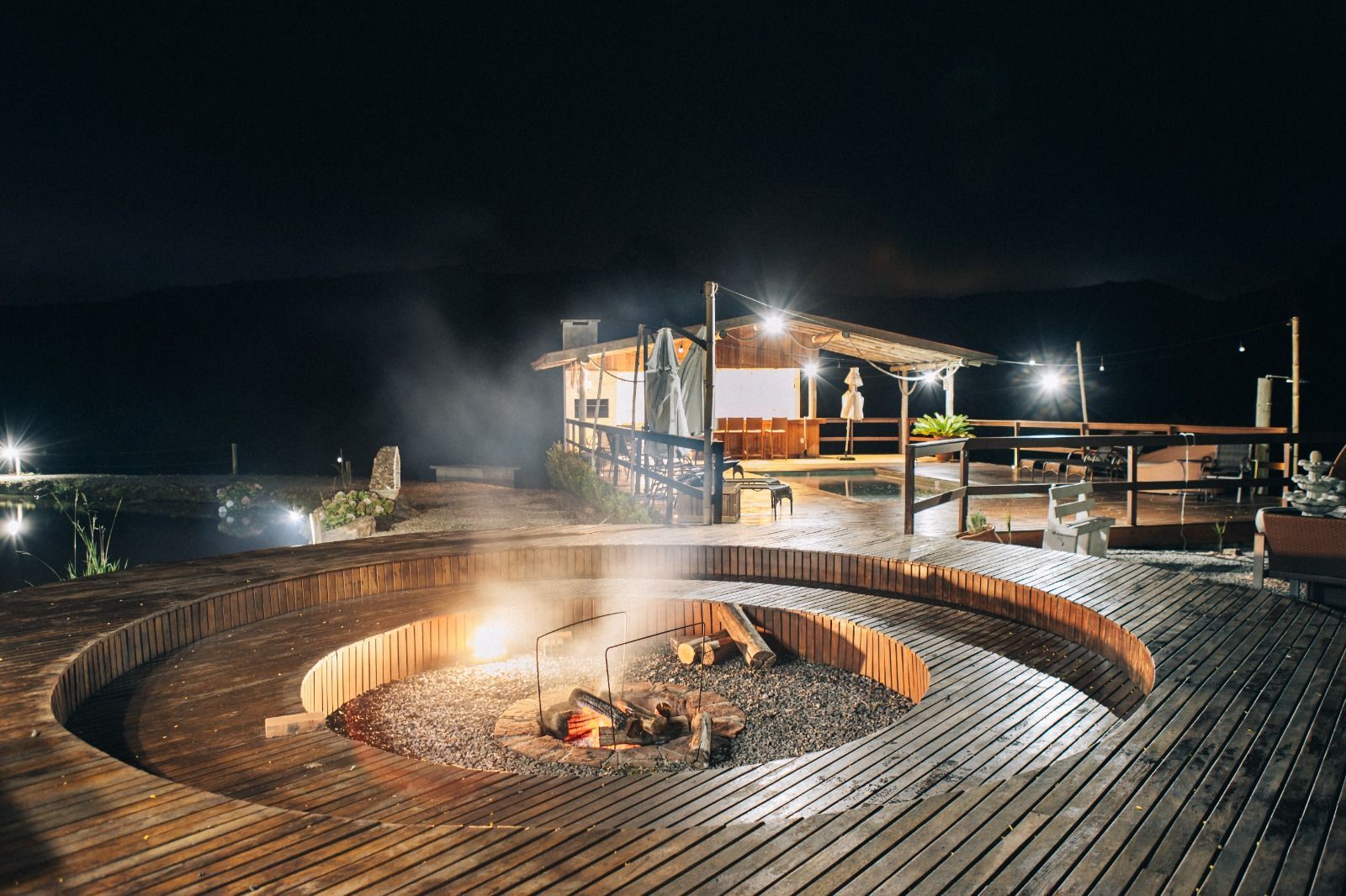Fogueira noturna à beira-mar em um deck de madeira com uma cabana iluminada ao fundo.