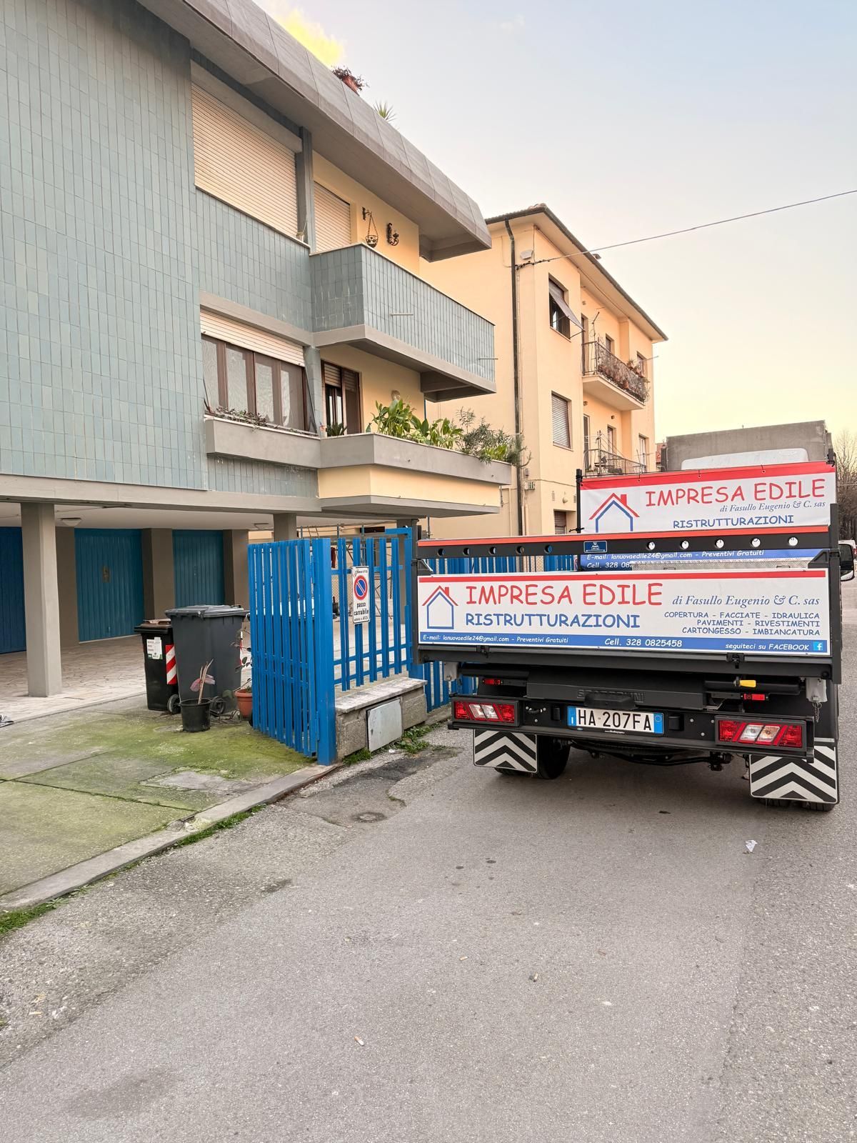 Operai con i gilet arancioni su un ascensore, vicino alla Torre Pendente di Pisa, intenti a pulire l'esterno di un edificio. Giornata di sole.